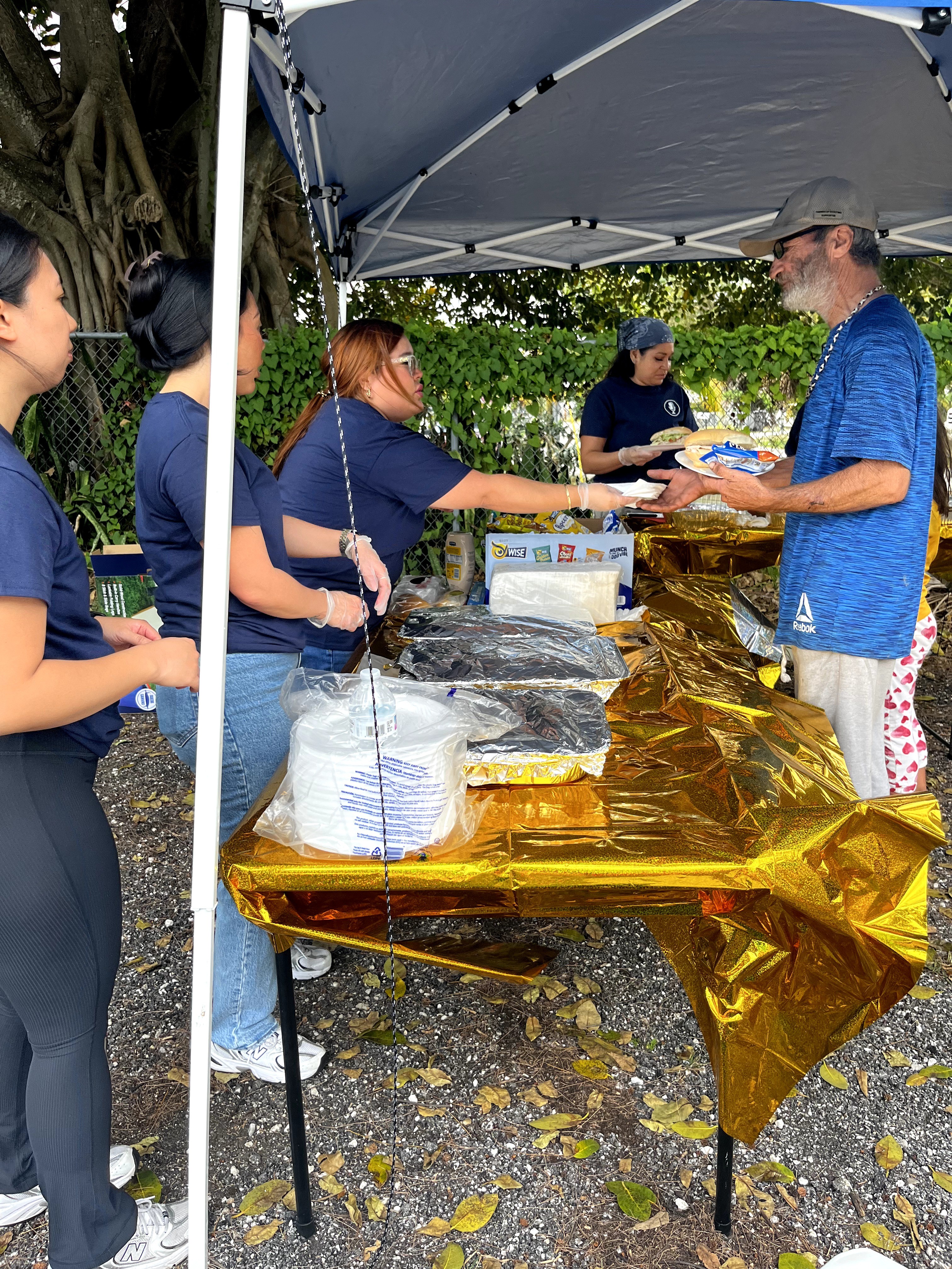 People in blue shirts are serving food at a table covered with gold foil under a canopy outdoors.