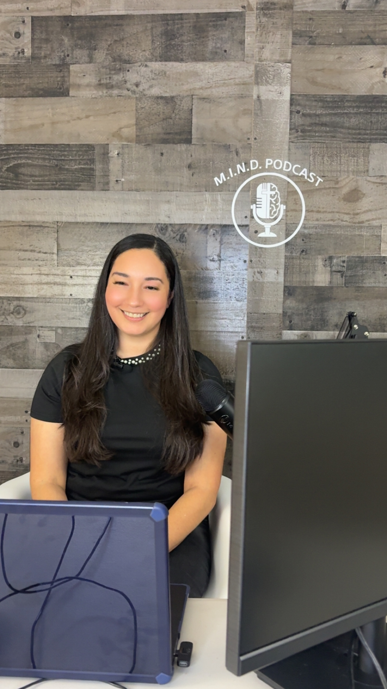 Young woman with long dark hair smiling, sitting in front of a wooden wall, with a computer monitor and microphone icon above her head.