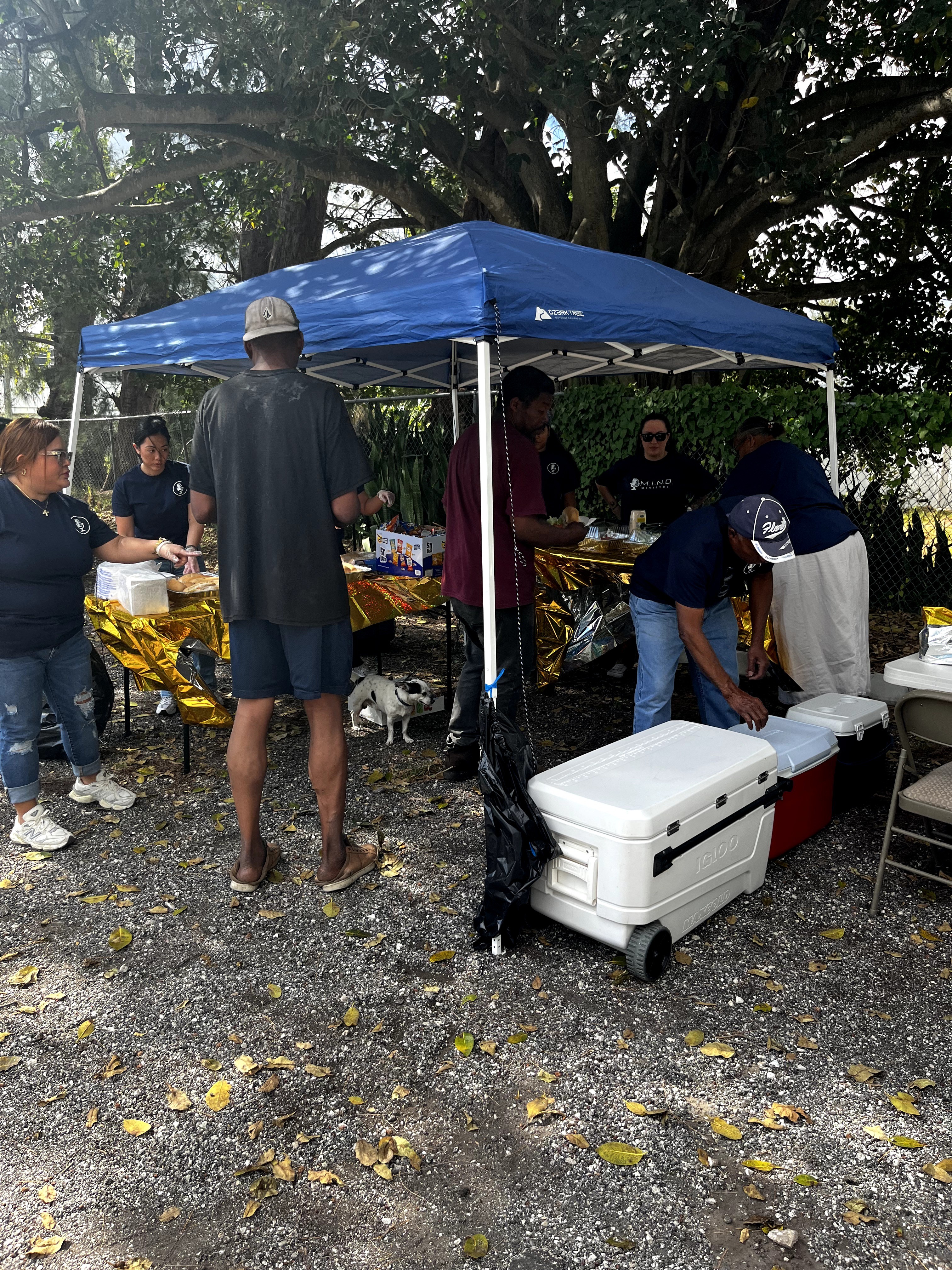 People gather around a blue canopy tent with tables and a cooler outdoors, trees in the background, gravel ground.