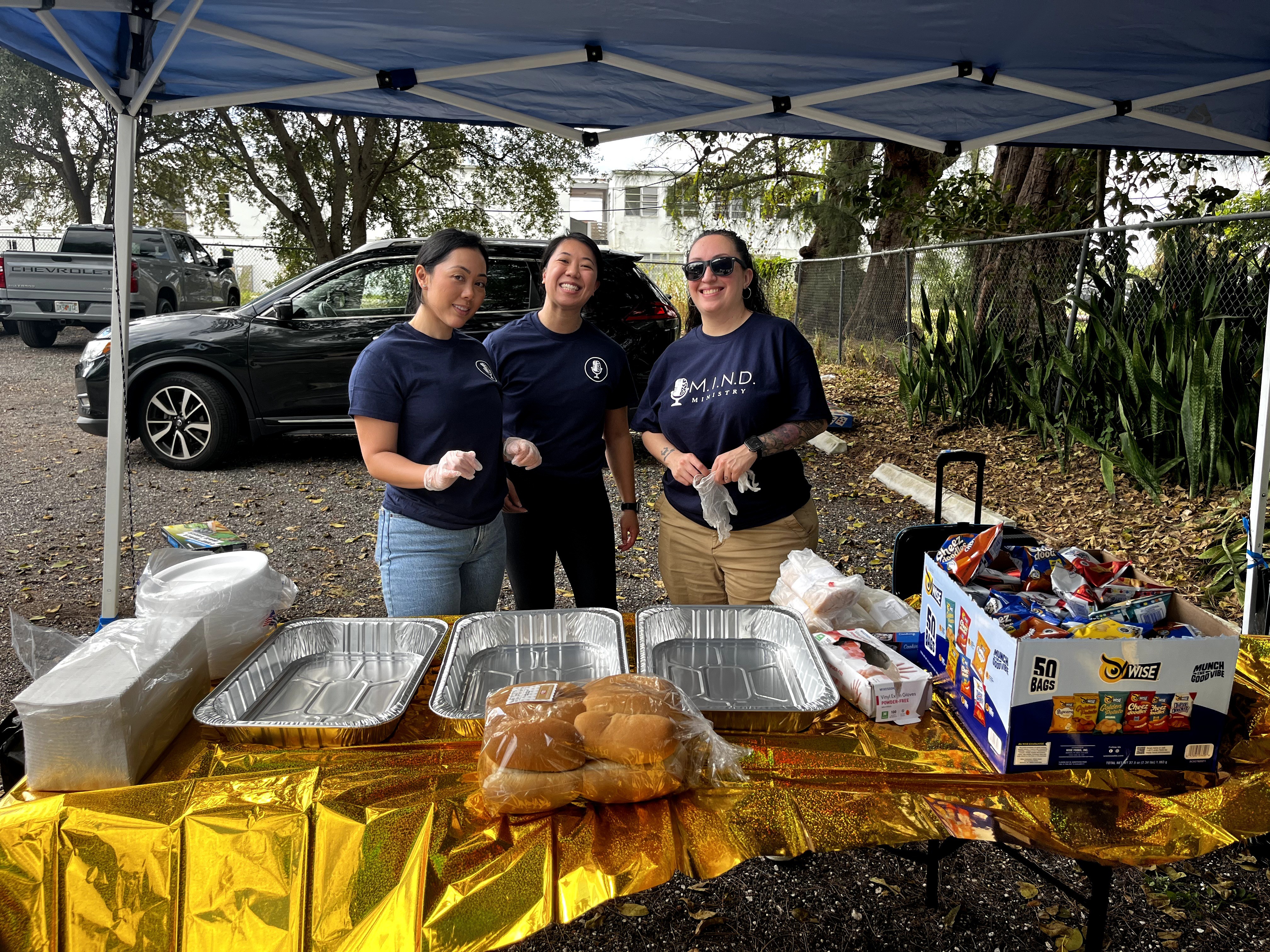 Three women standing behind a table with food under a blue canopy outdoors, with trees and a car in the background.
