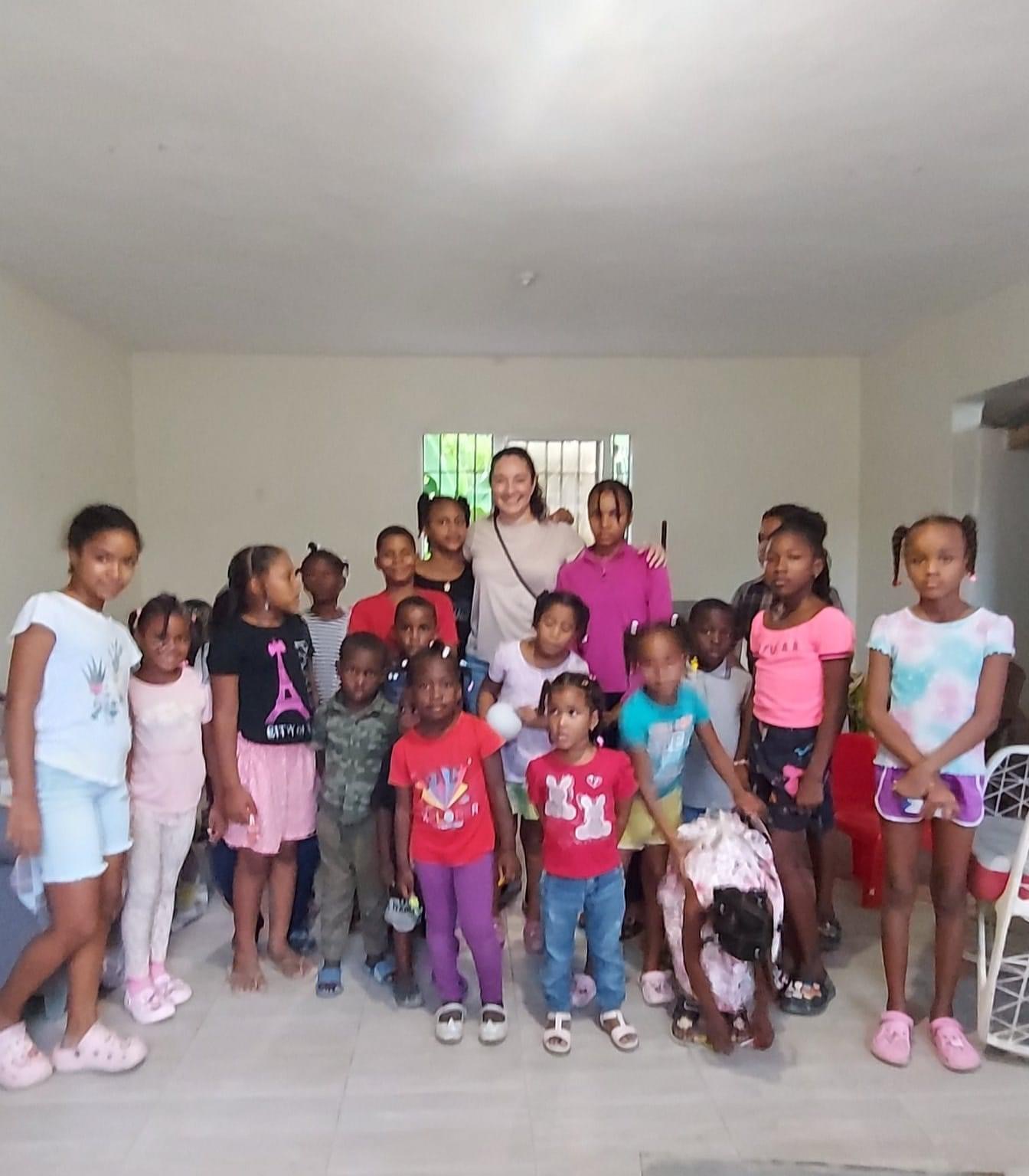 Group of children and an adult standing indoors in front of a window, smiling, with some children holding bags or backpacks.