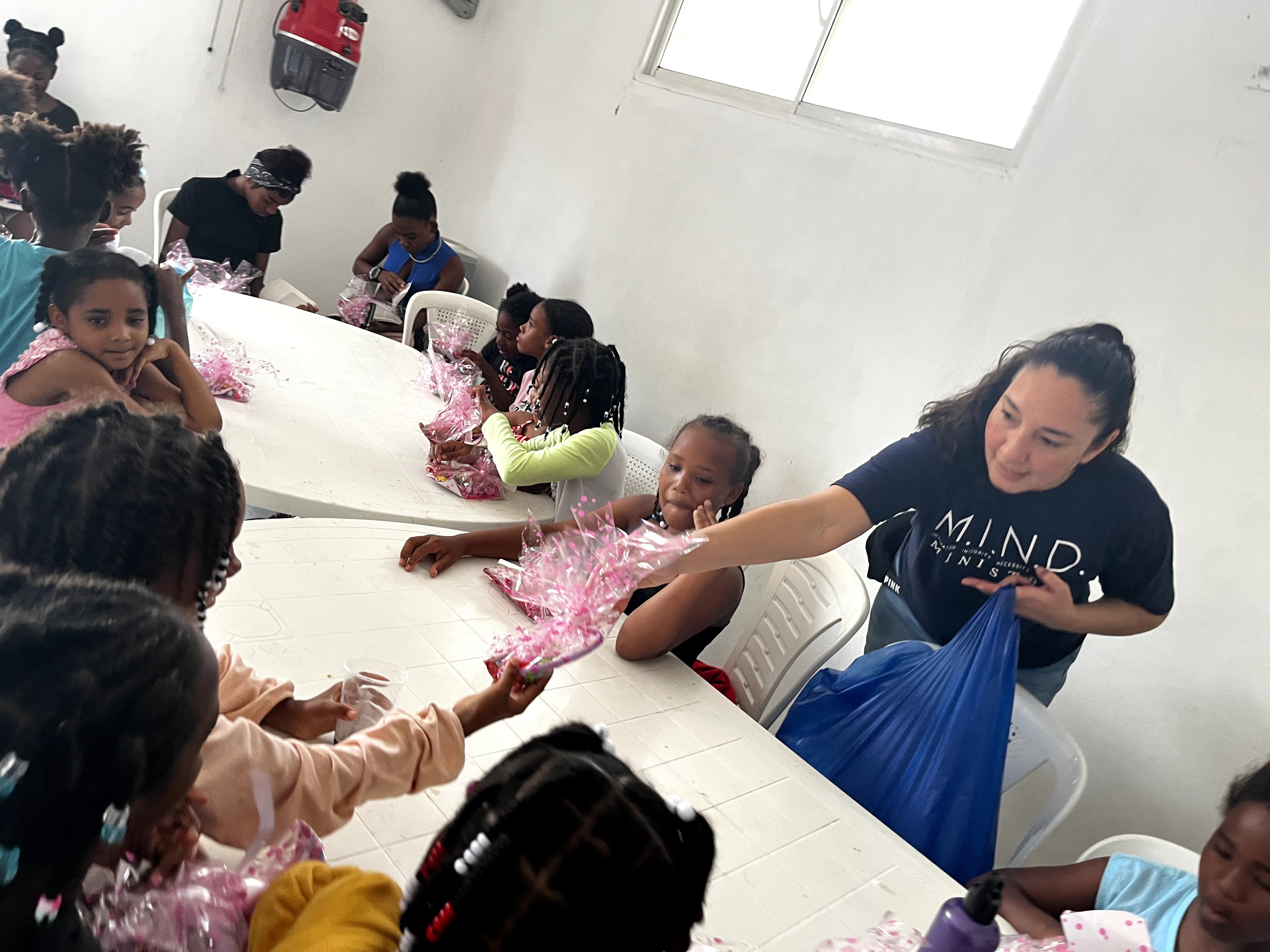Children and a woman sit around a table, with the woman handing out pink items, in a bright room.