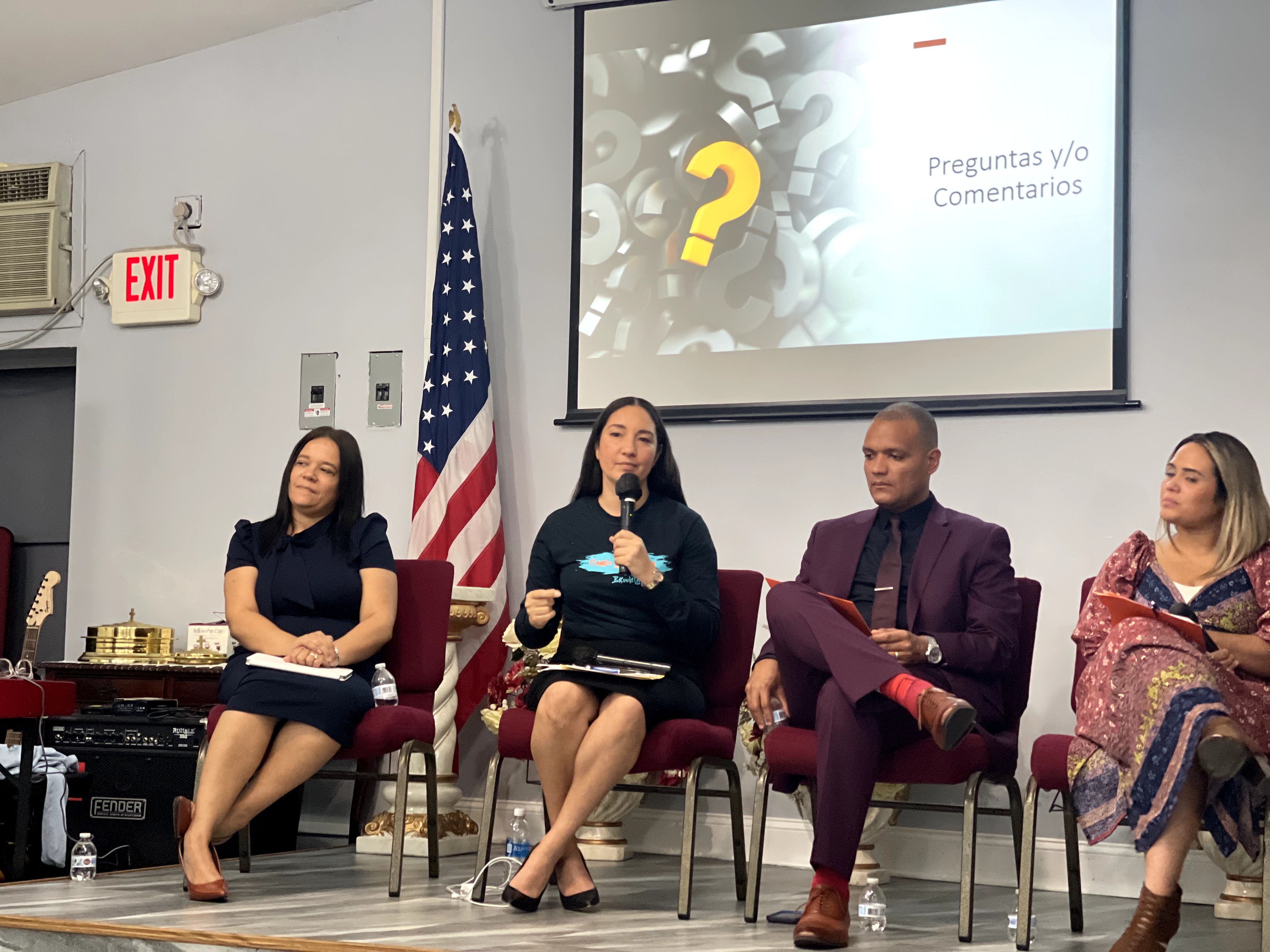 Four people seated on a panel in a room with an American flag and a presentation screen behind them.