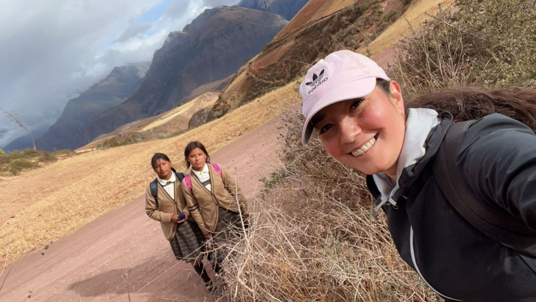 Three people standing on a dirt path in a mountainous landscape, smiling, with mountains and cloudy sky in background.