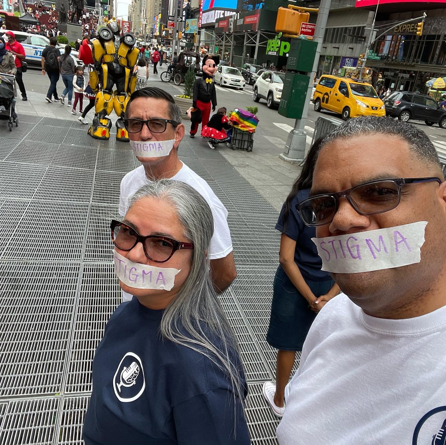 Three people with paper masks labeled 'STIGMA' on their mouths, standing on a busy city sidewalk.