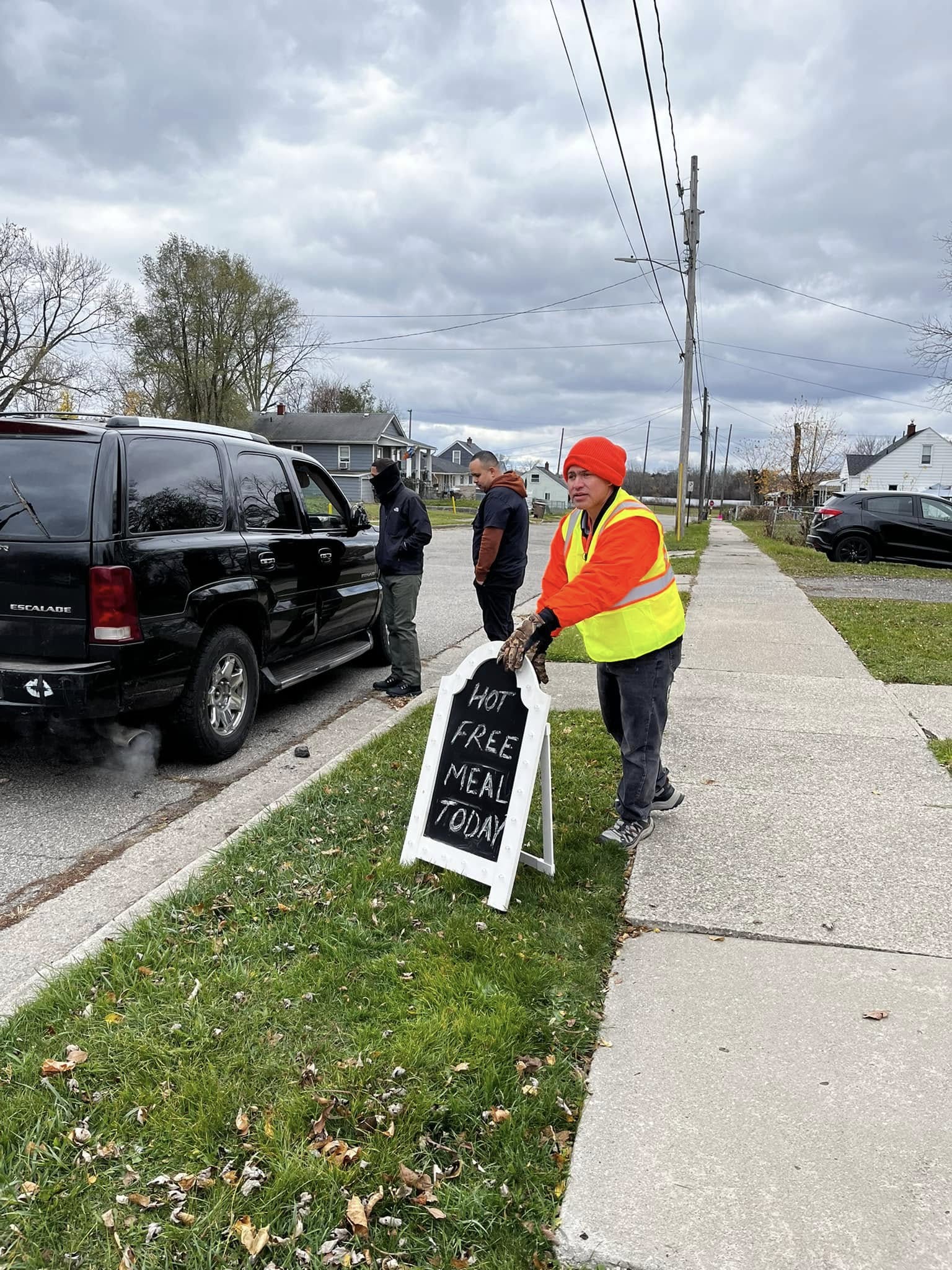 Person in a red hat and yellow vest standing next to a black vehicle on a sidewalk, holding a sign about free meals.