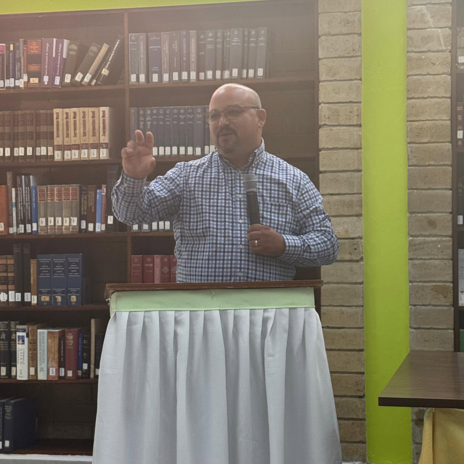 Man speaking at a podium in a library, holding a microphone, with bookshelves in the background.