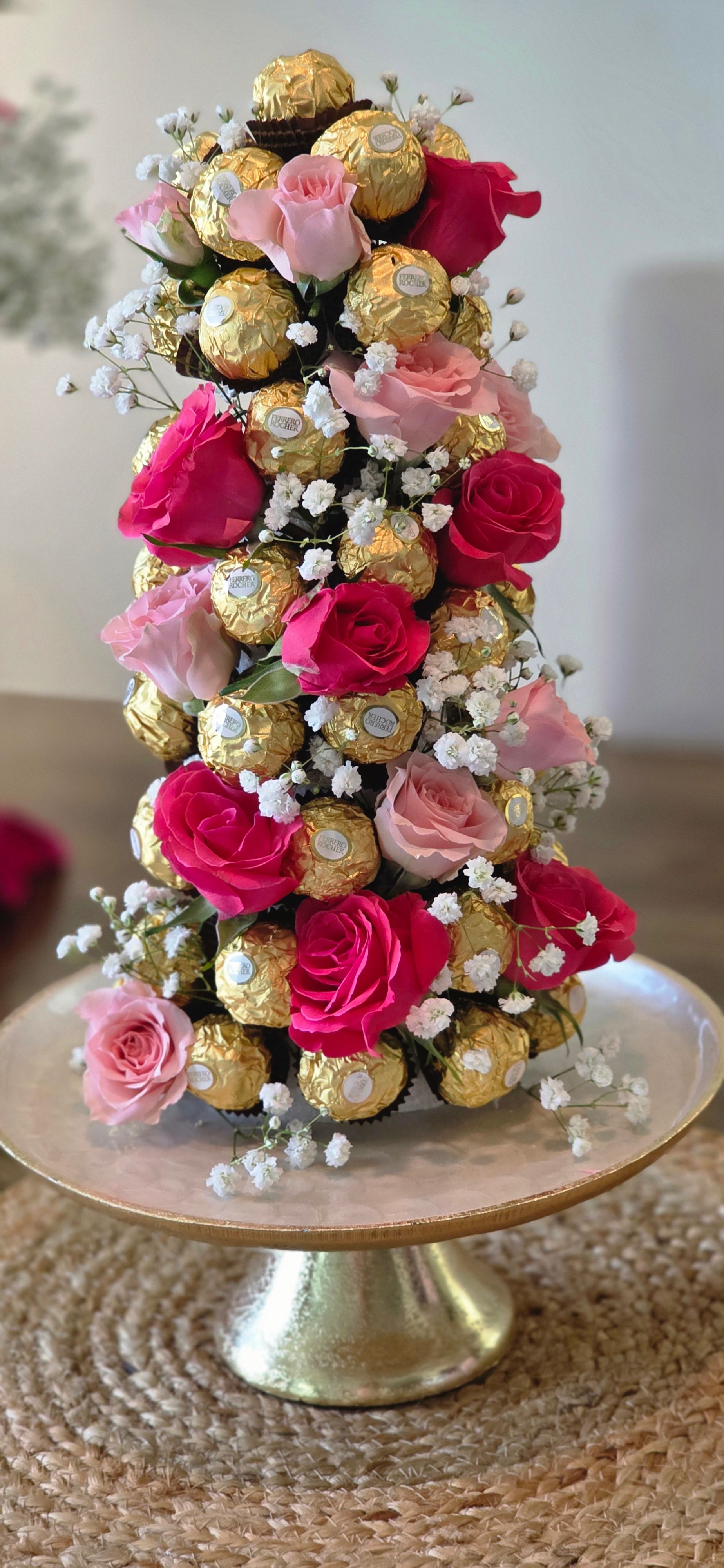 Flower arrangement with pink and red roses, gold-wrapped chocolates, and white baby's breath on a round table.