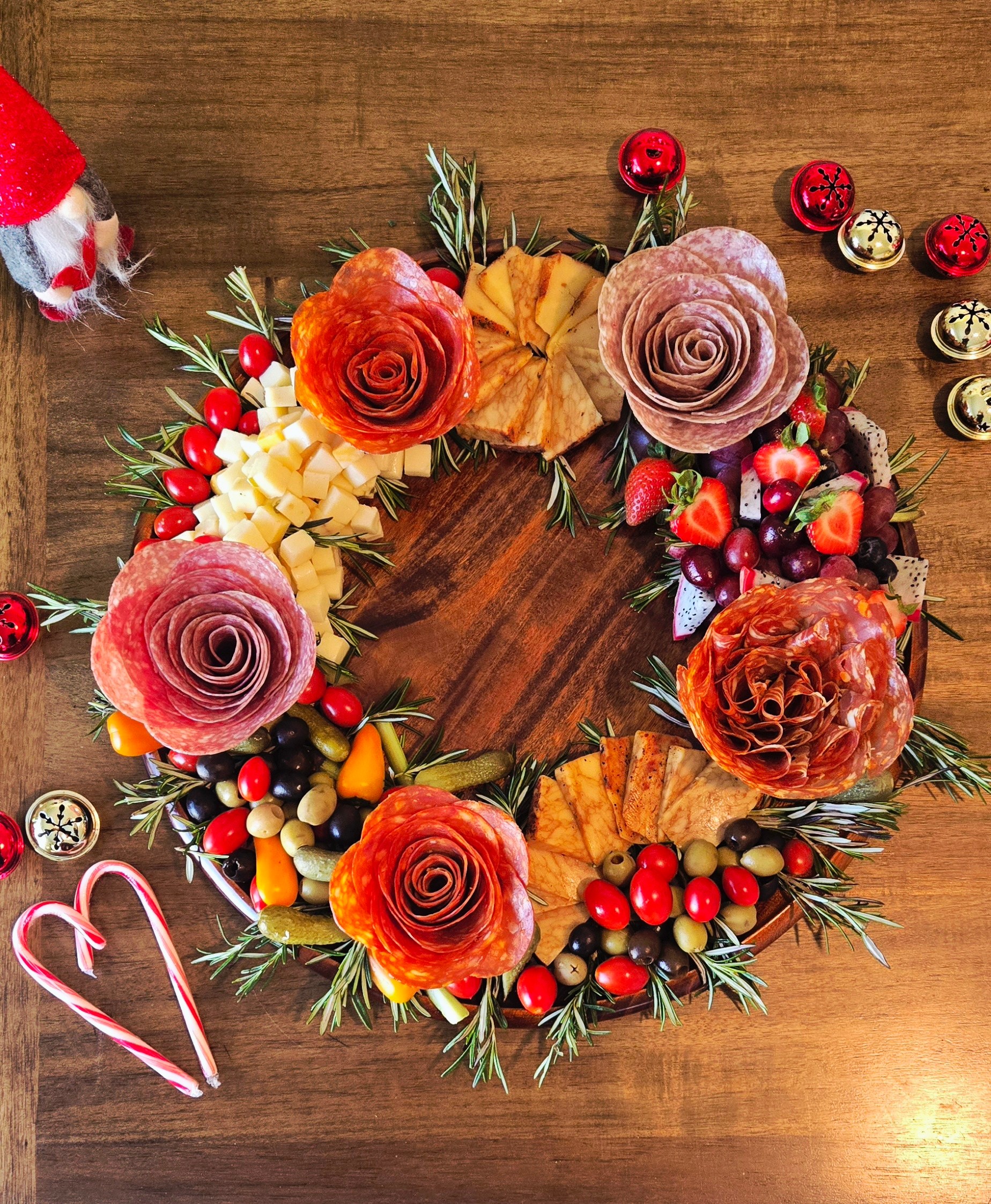 Decorative wreath with roses, berries, leaves, and pine branches on a wooden surface, with small bells and a candy cane nearby.