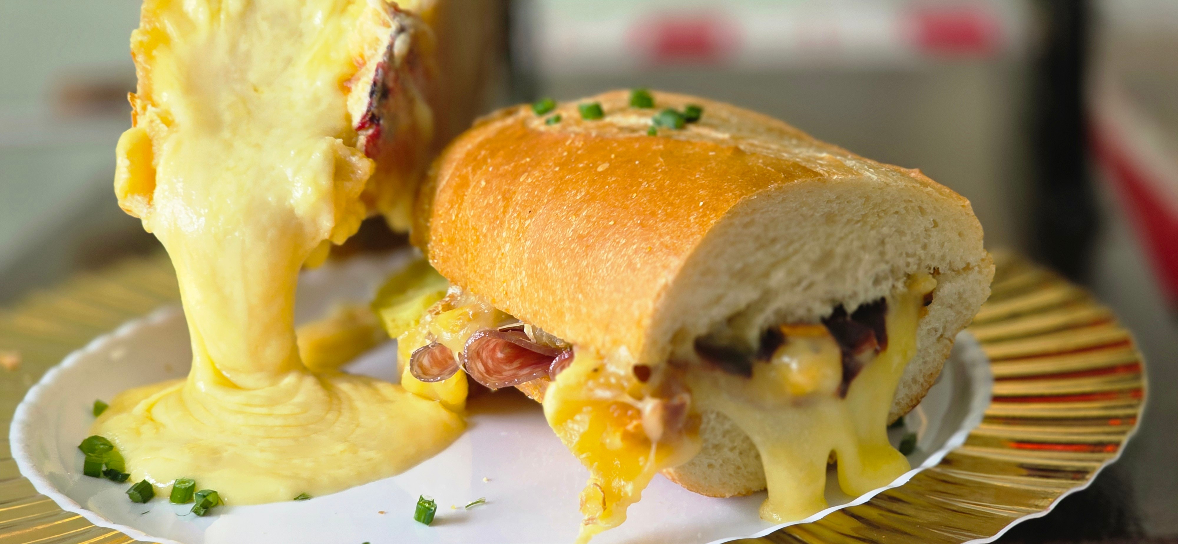 Sandwich with melted cheese, meat, and vegetables on a bun, with cheese stretching out, on a plate.
