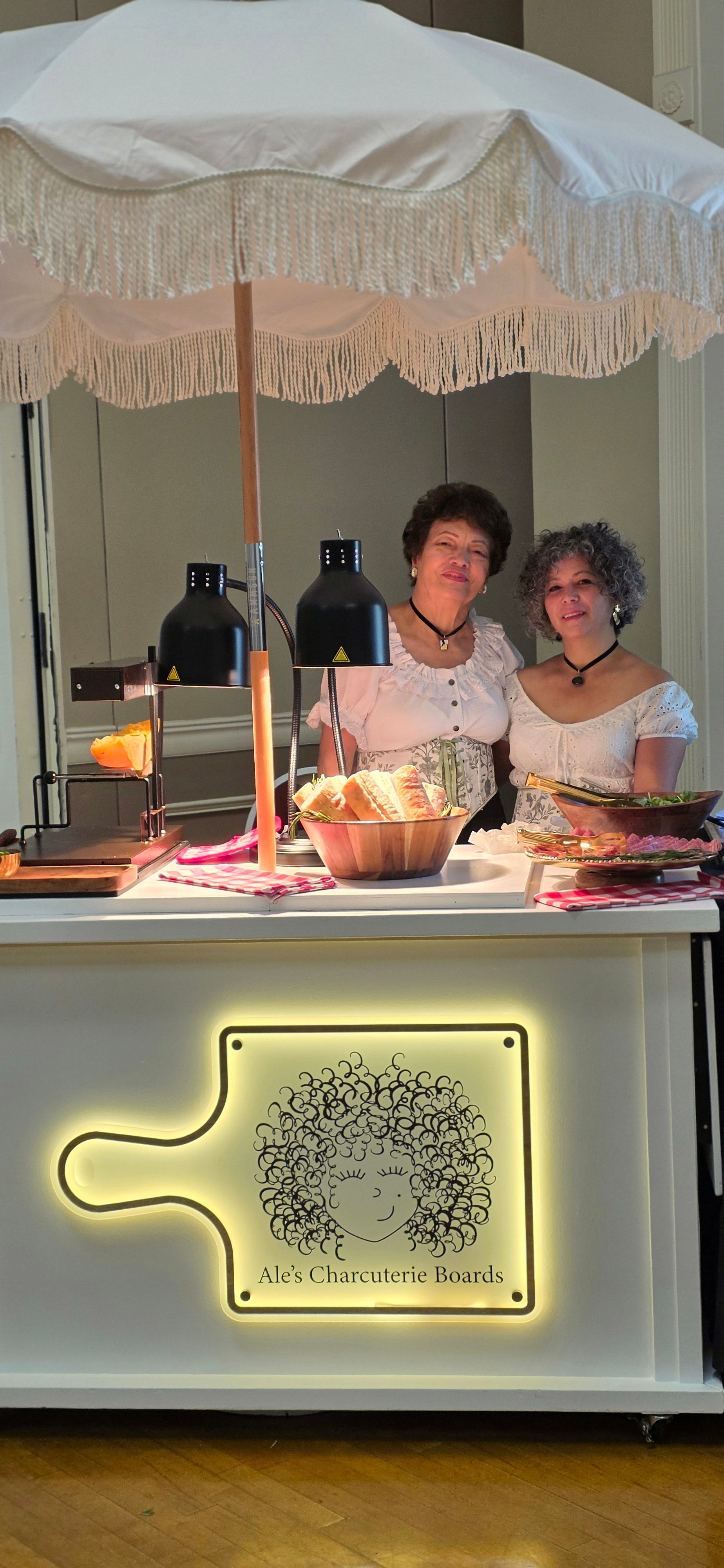 Two women sit at a table with drinks, in a room with a decorative wall sign, seen from the side.