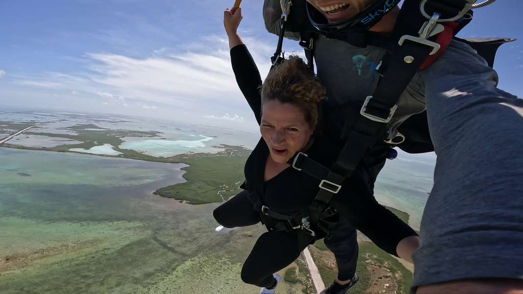 Person and child tandem skydiving over a landscape with water and land, child appears excited, sky is partly cloudy.