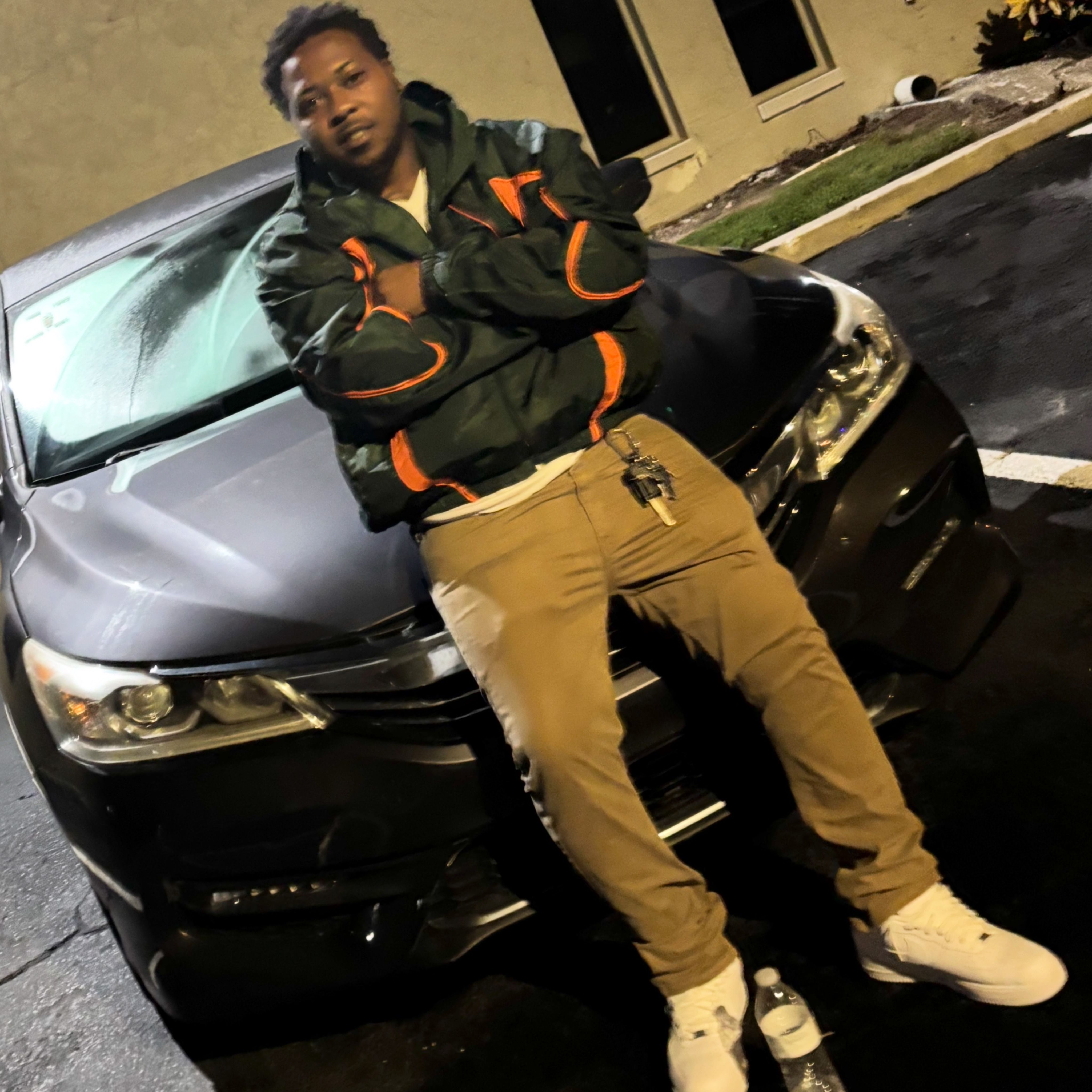 Person lying on a car at night, wearing a black jacket with orange accents, tan pants, and white shoes.