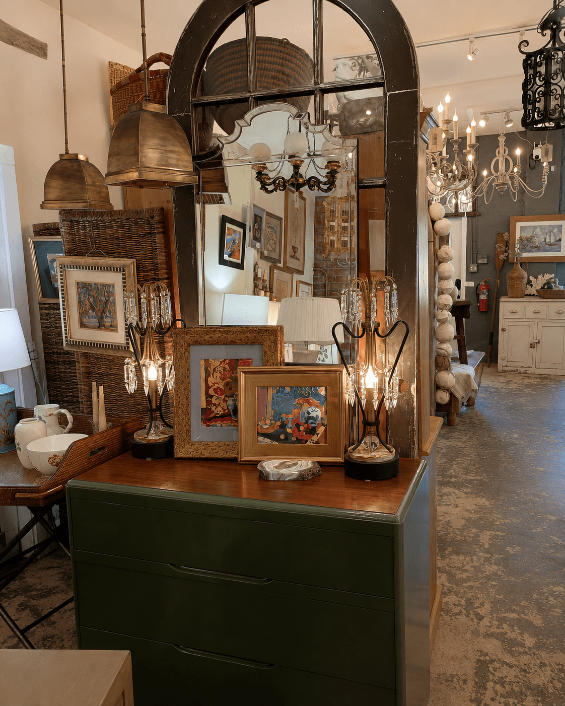 Wooden dresser with framed pictures, lamps, and decorative items, reflected in a large mirror, in an antique store interior.
