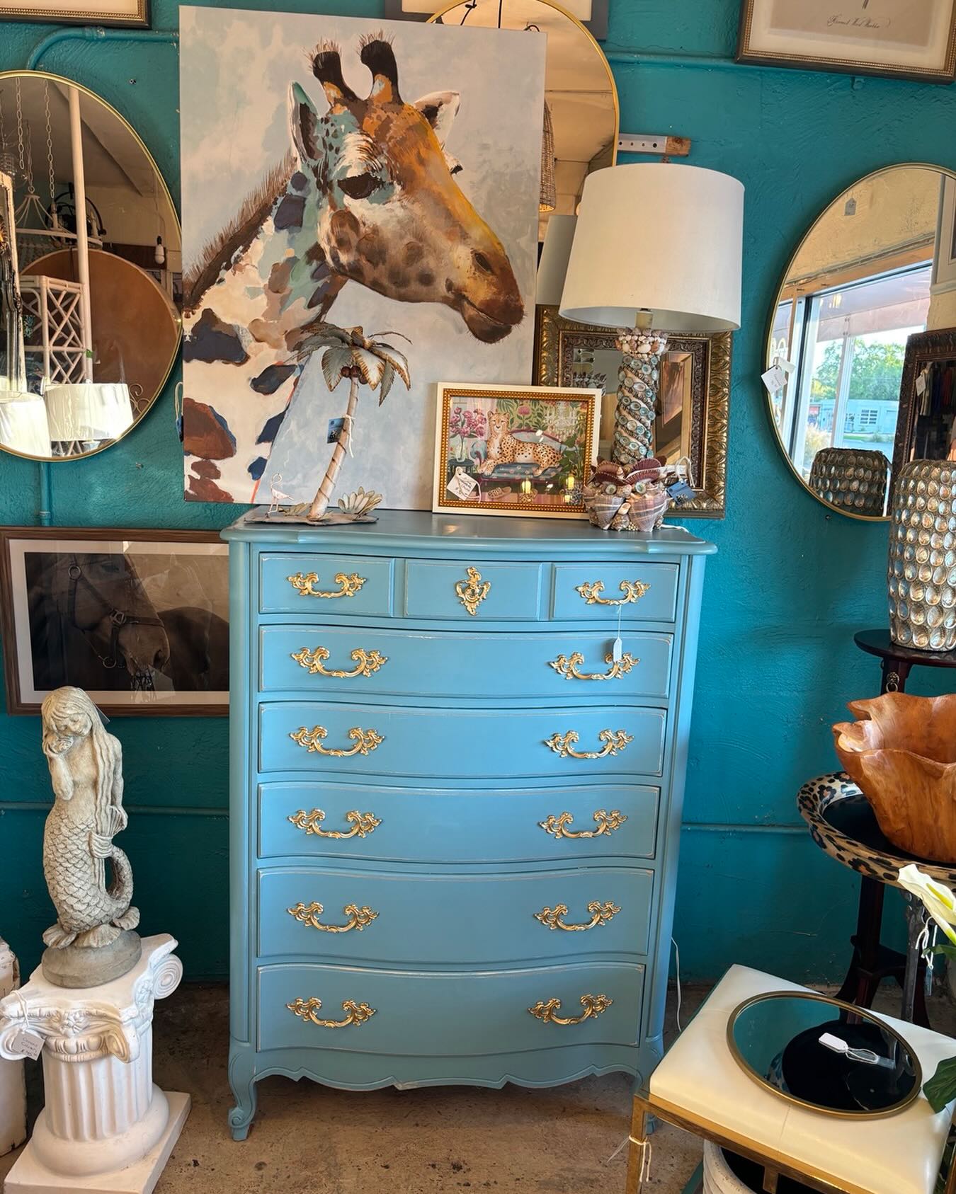 Blue dresser with gold handles, artwork of a giraffe, lamp, and framed picture on top, in a room with mirrors and decor.