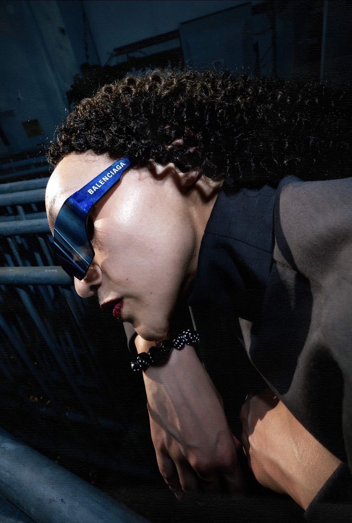 Person with curly hair wearing glasses and a dark outfit, looking downward, in a dimly lit environment.