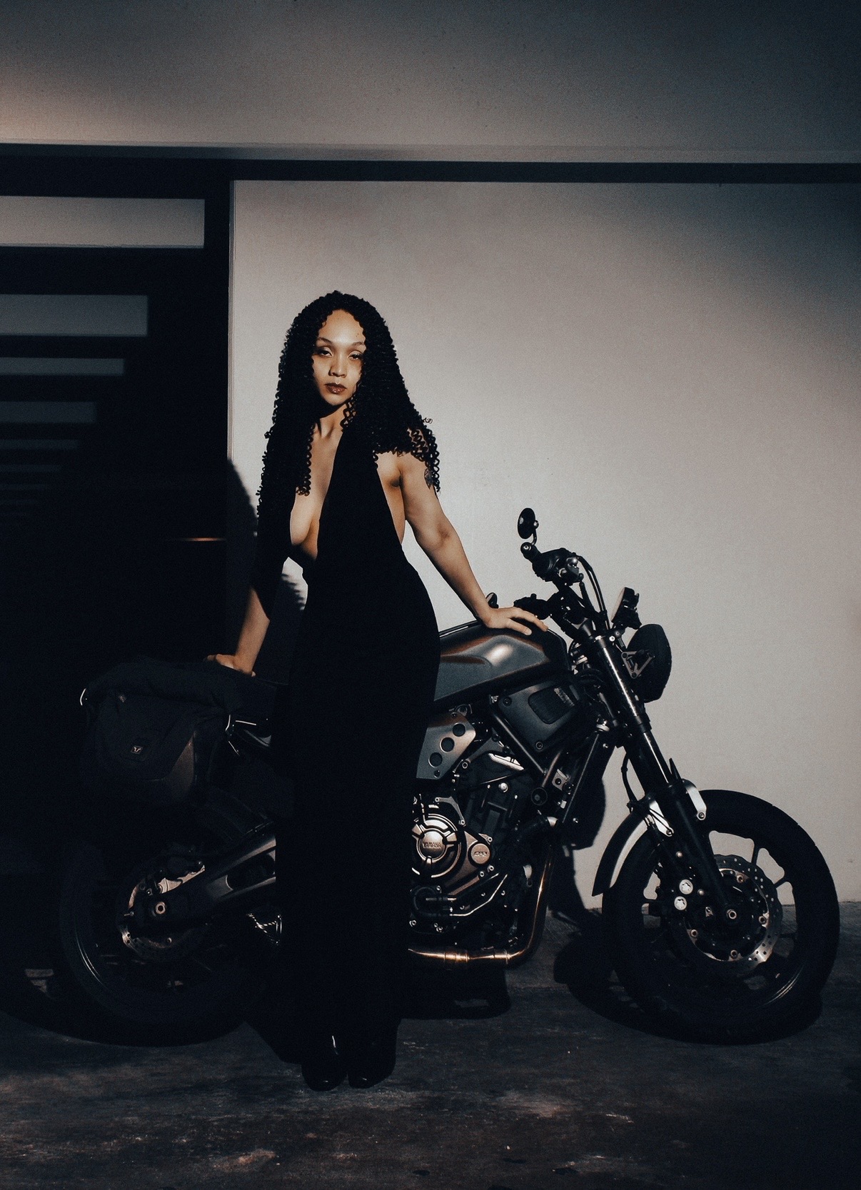 Woman with long curly hair in black outfit standing next to a motorcycle in a dimly lit room.