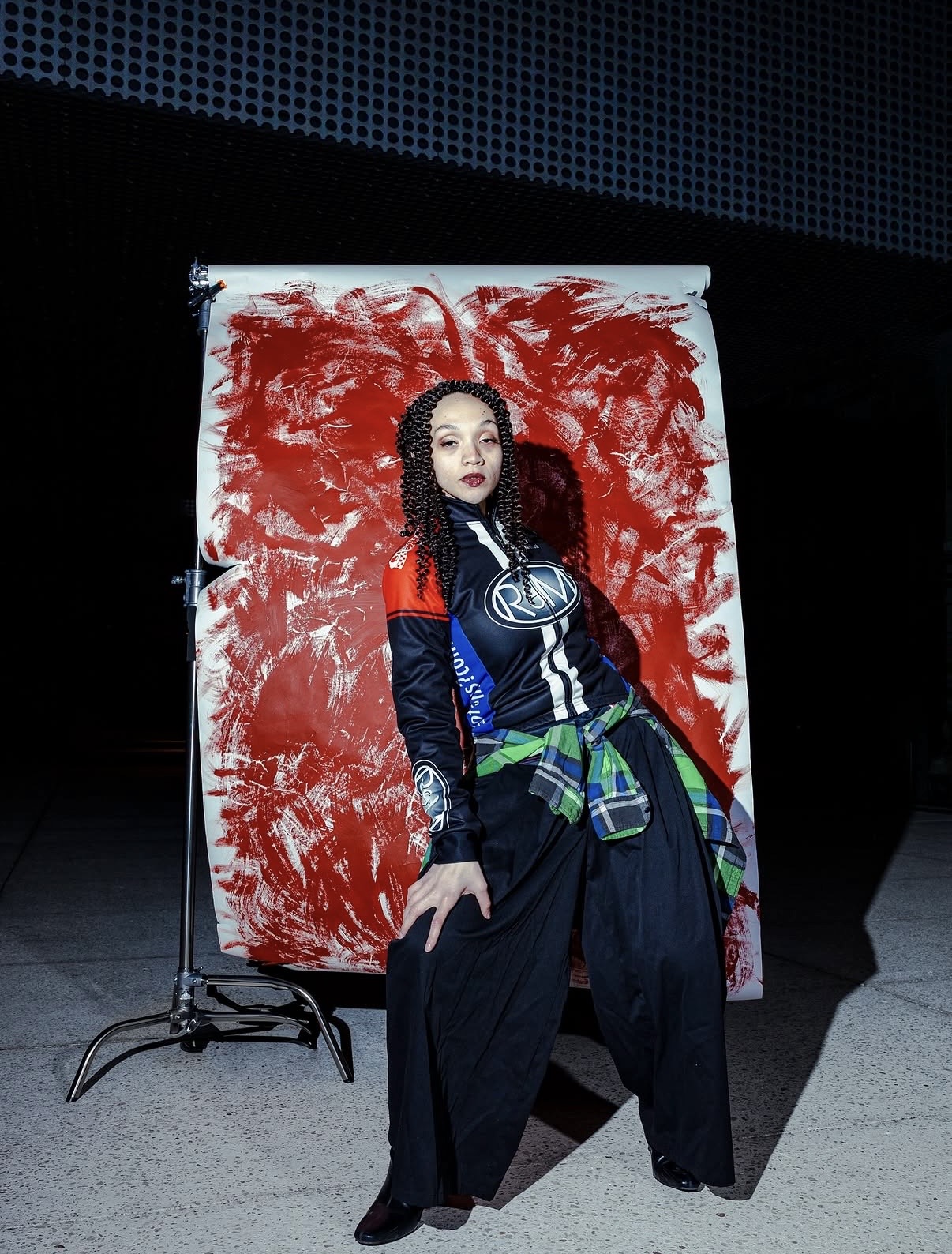 Young woman with curly hair and makeup, standing in front of a red and white abstract background, wearing a colorful jacket and wide pants.