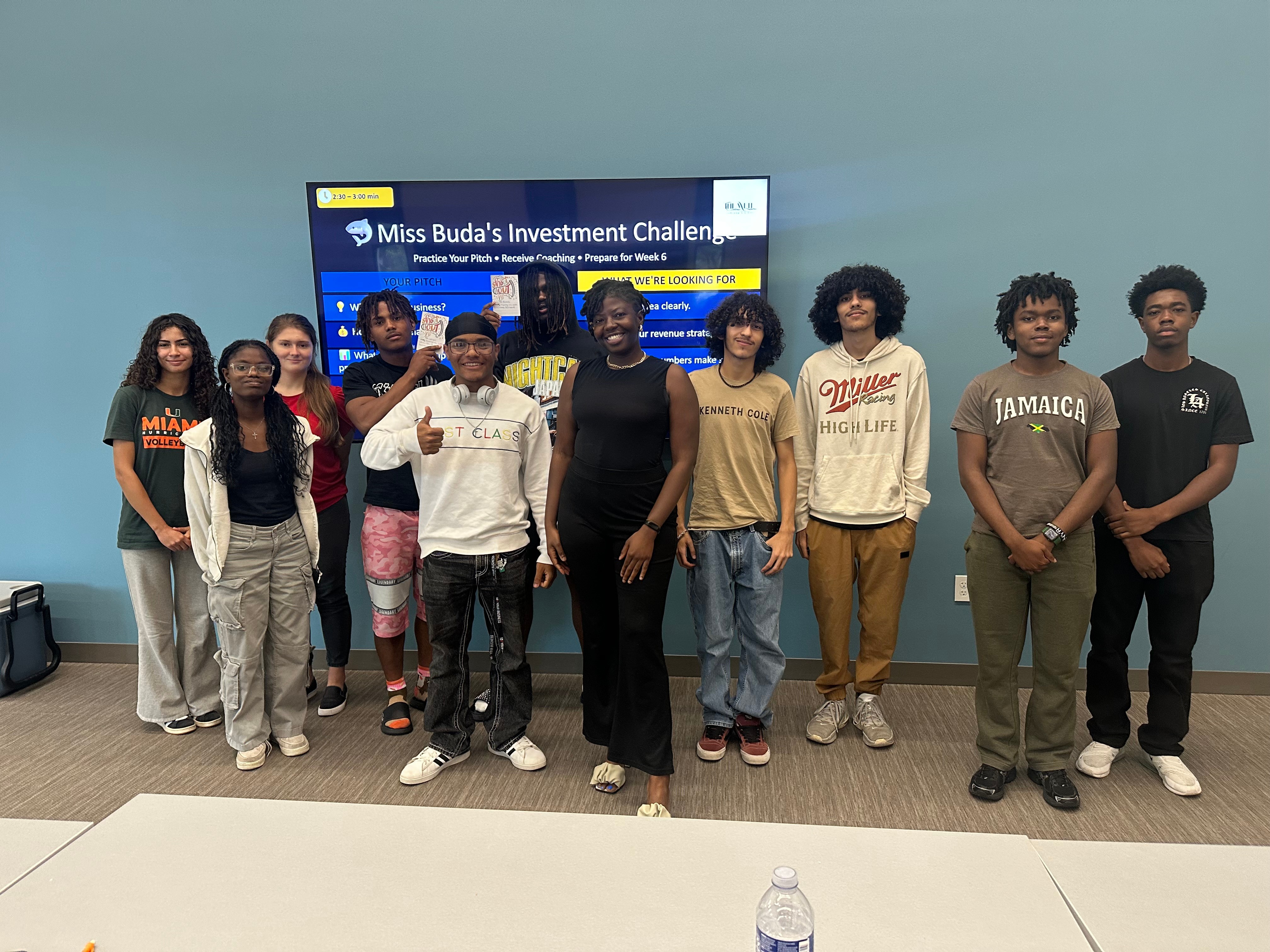 Group of ten young people standing in front of a screen, smiling, indoors. The screen displays 'Miss Buda's Investment Challenge.'