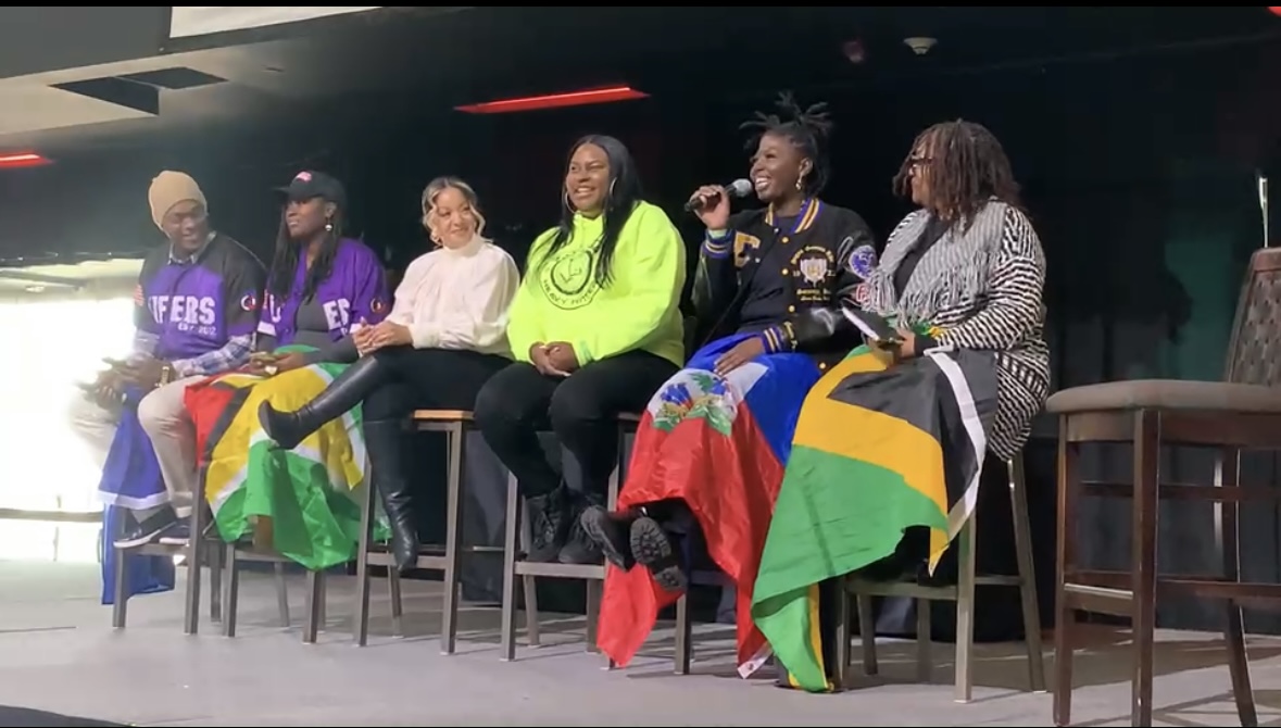 Six women sit on a stage, some holding flags, with a dark background and red lighting above, smiling and talking.