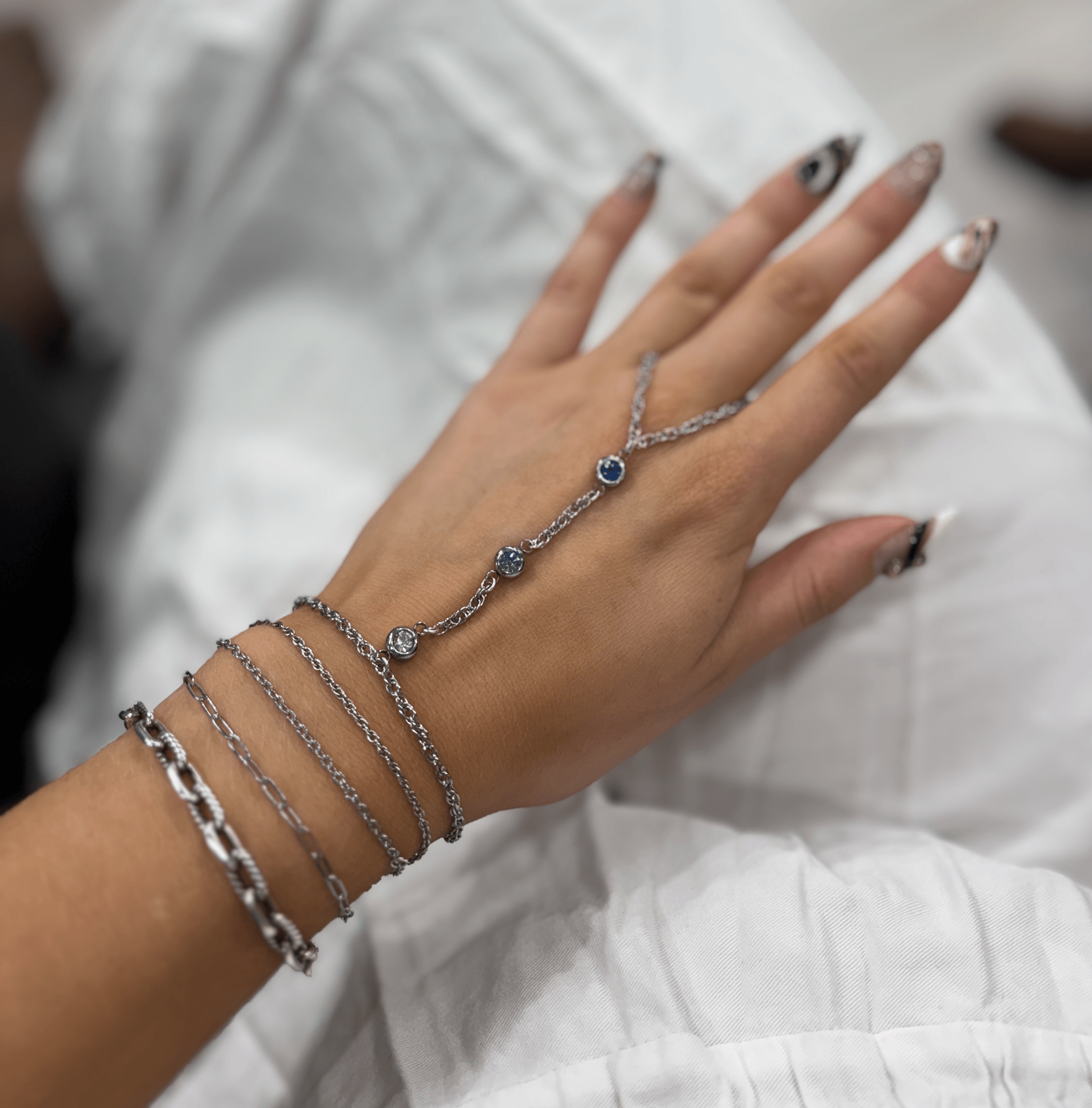 Hand with jewelry resting on white fabric, showing rings and bracelets, with painted nails. Word count: 20.