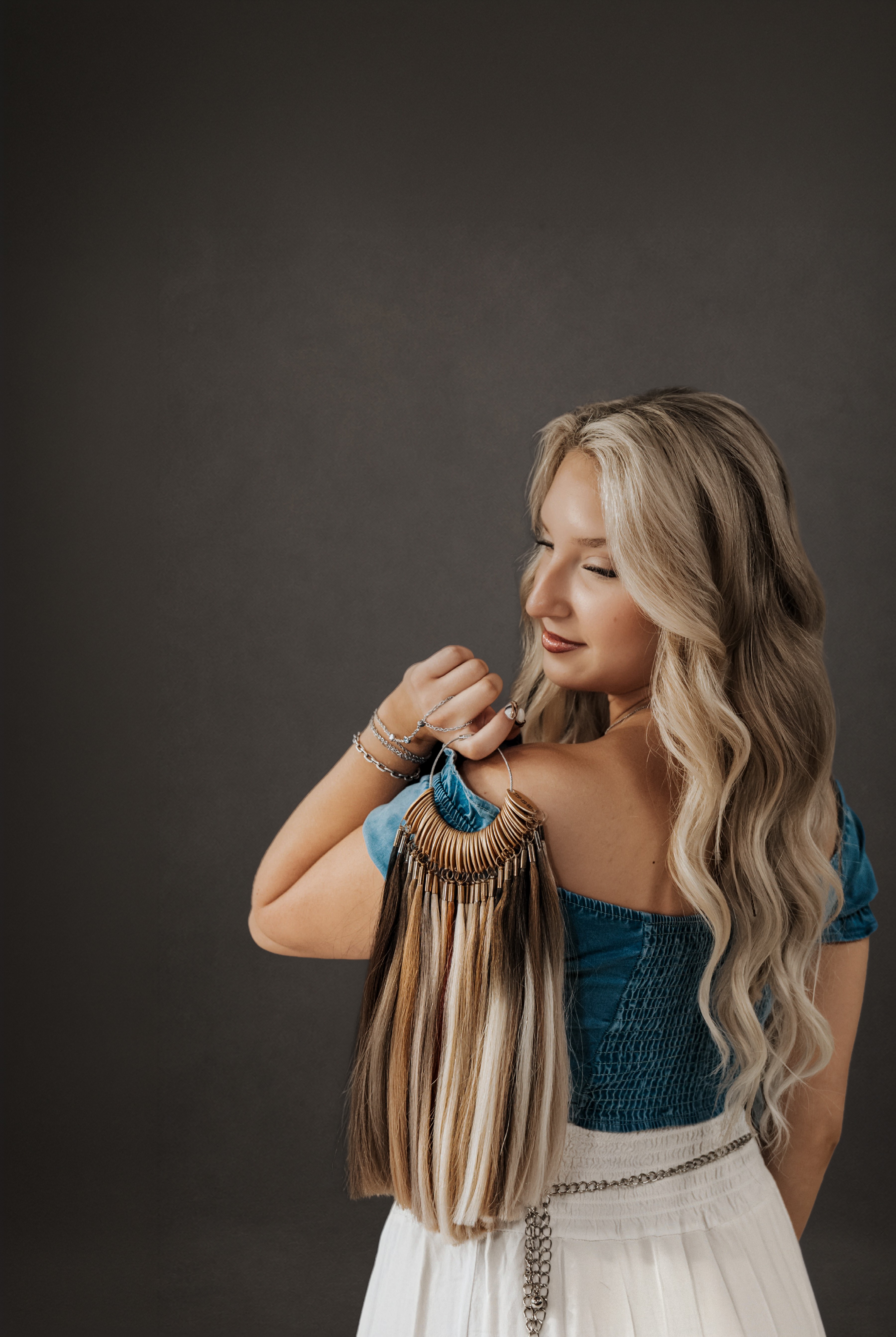 Woman with long, wavy blonde hair wearing a blue top and white skirt, holding her hair, facing sideways.