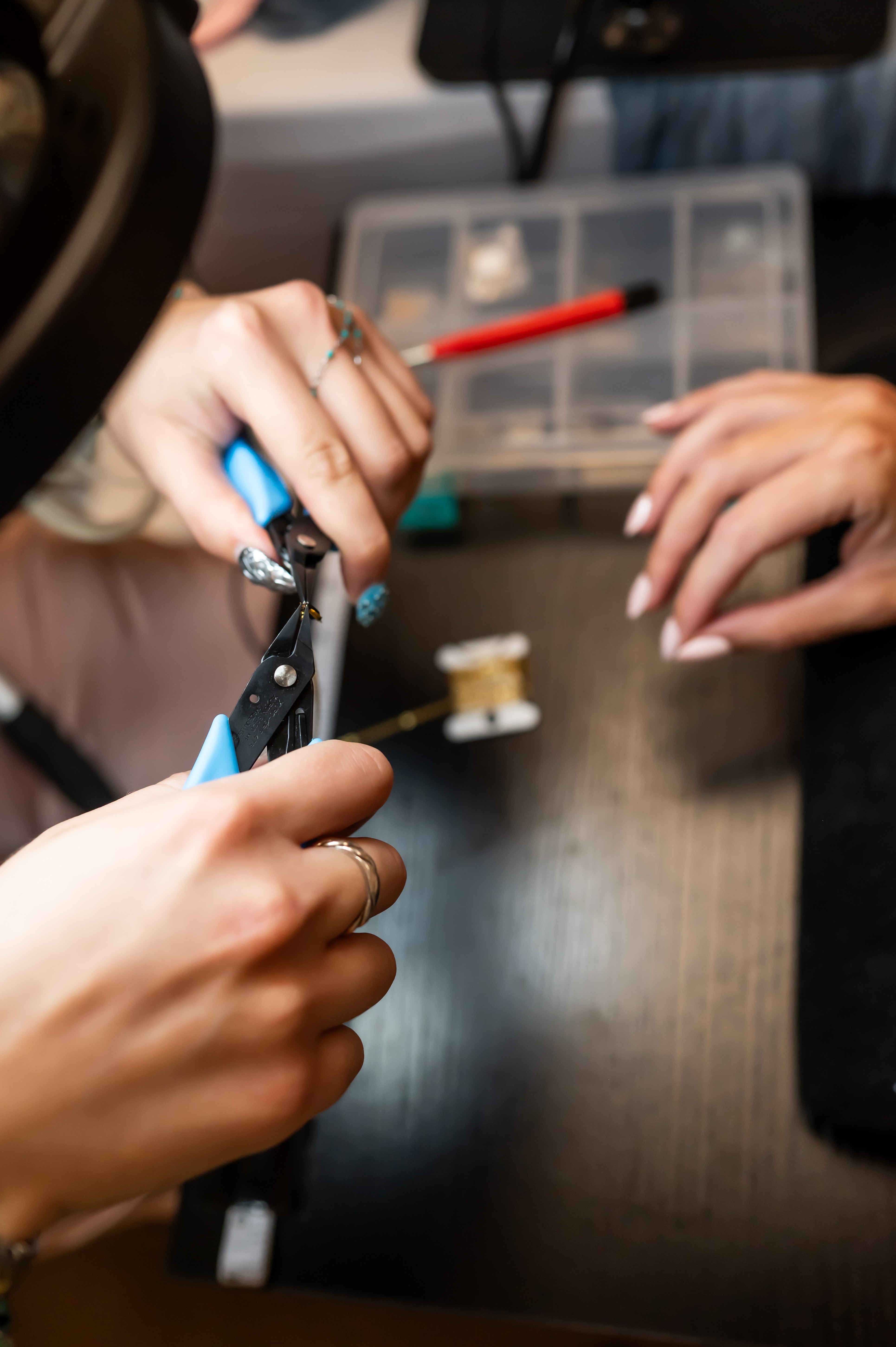 Hands working with jewelry tools over a dark table, with a plastic container in the background.
