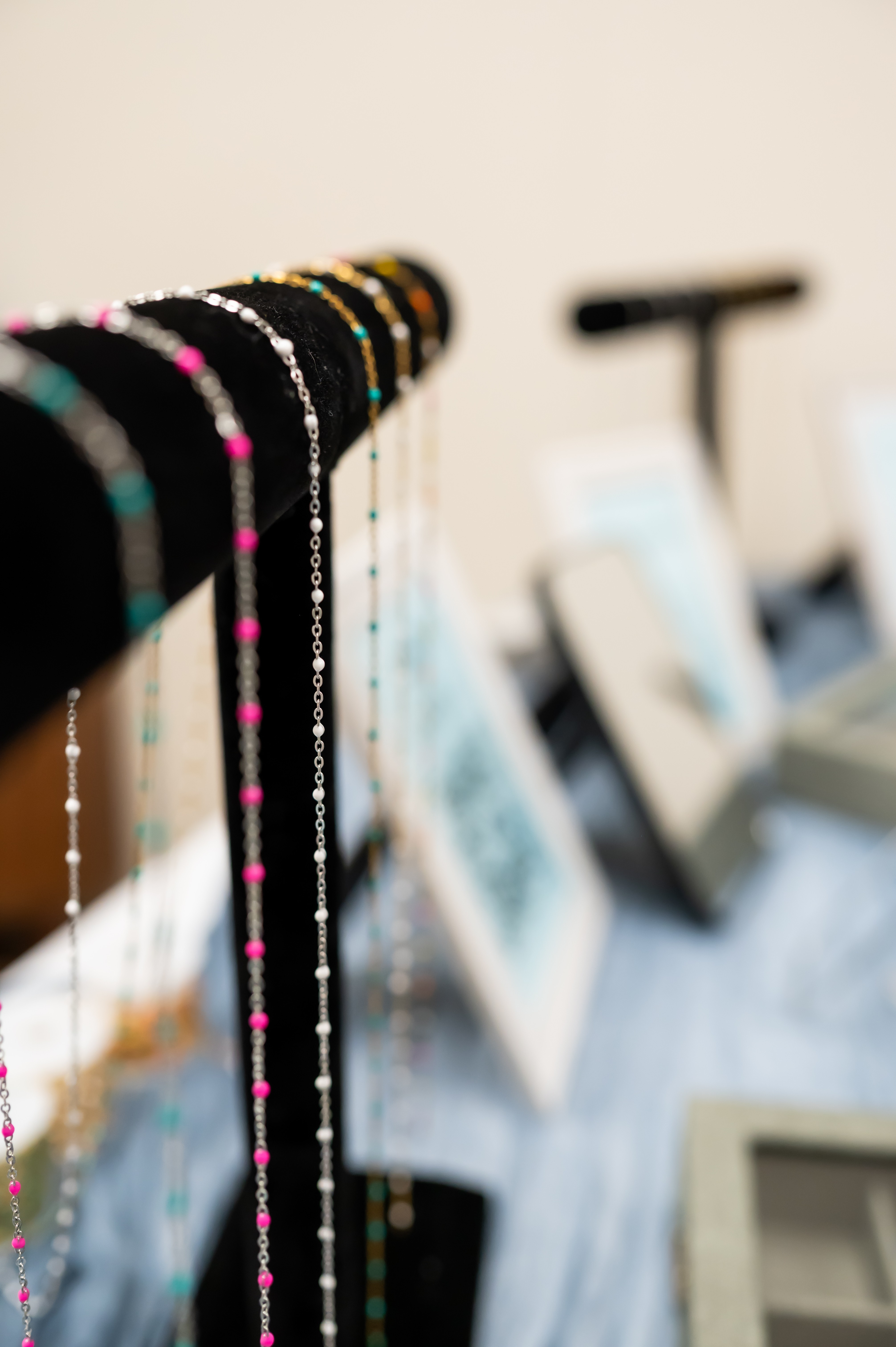 Display of colorful beaded necklaces on a black stand with blurred electronic devices in the background.