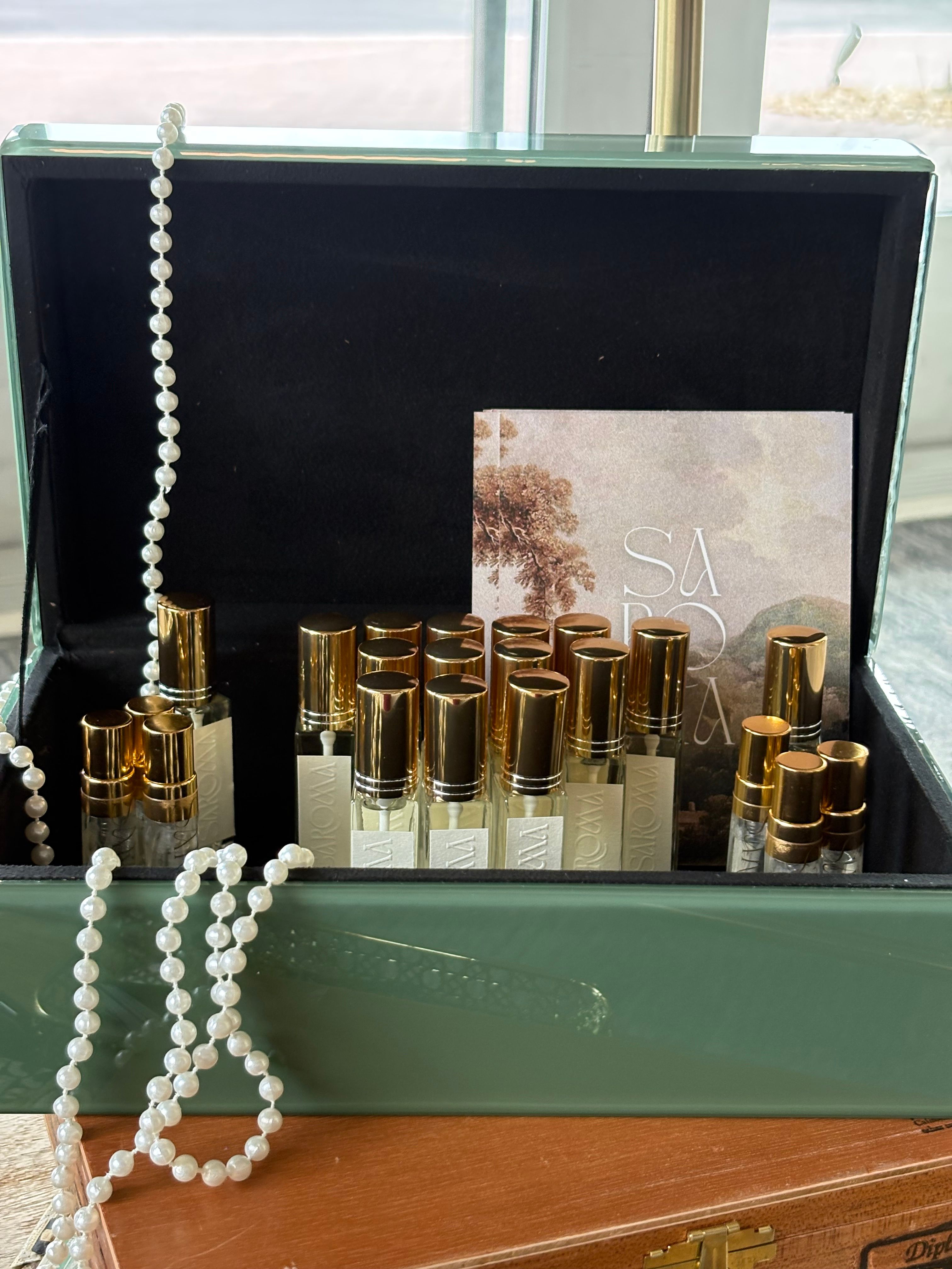 Open green box with gold bottles and a pearl necklace inside, on a wooden surface, with a window in the background.
