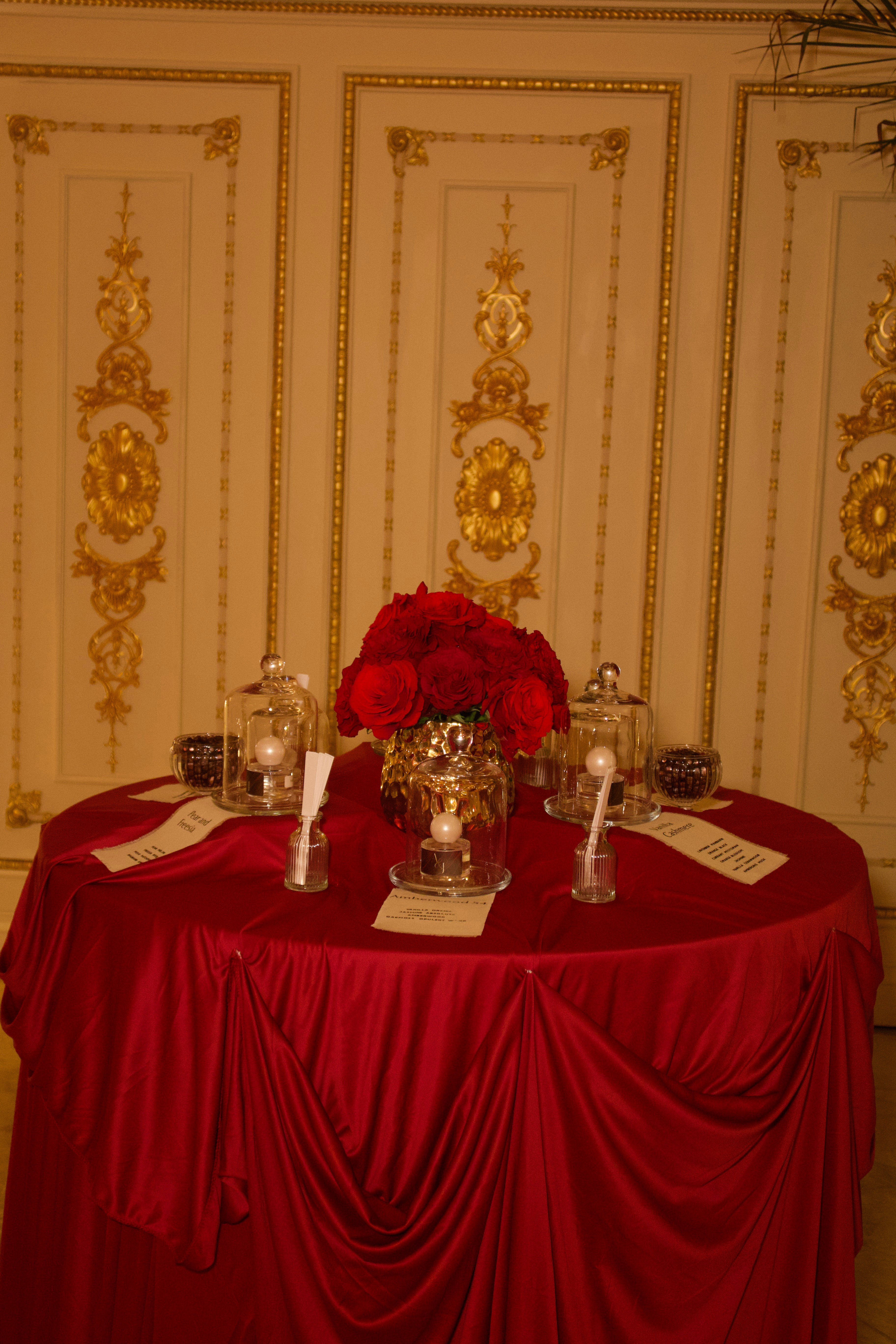Round table with red tablecloth, floral centerpiece, candles, and place settings in an ornate room.