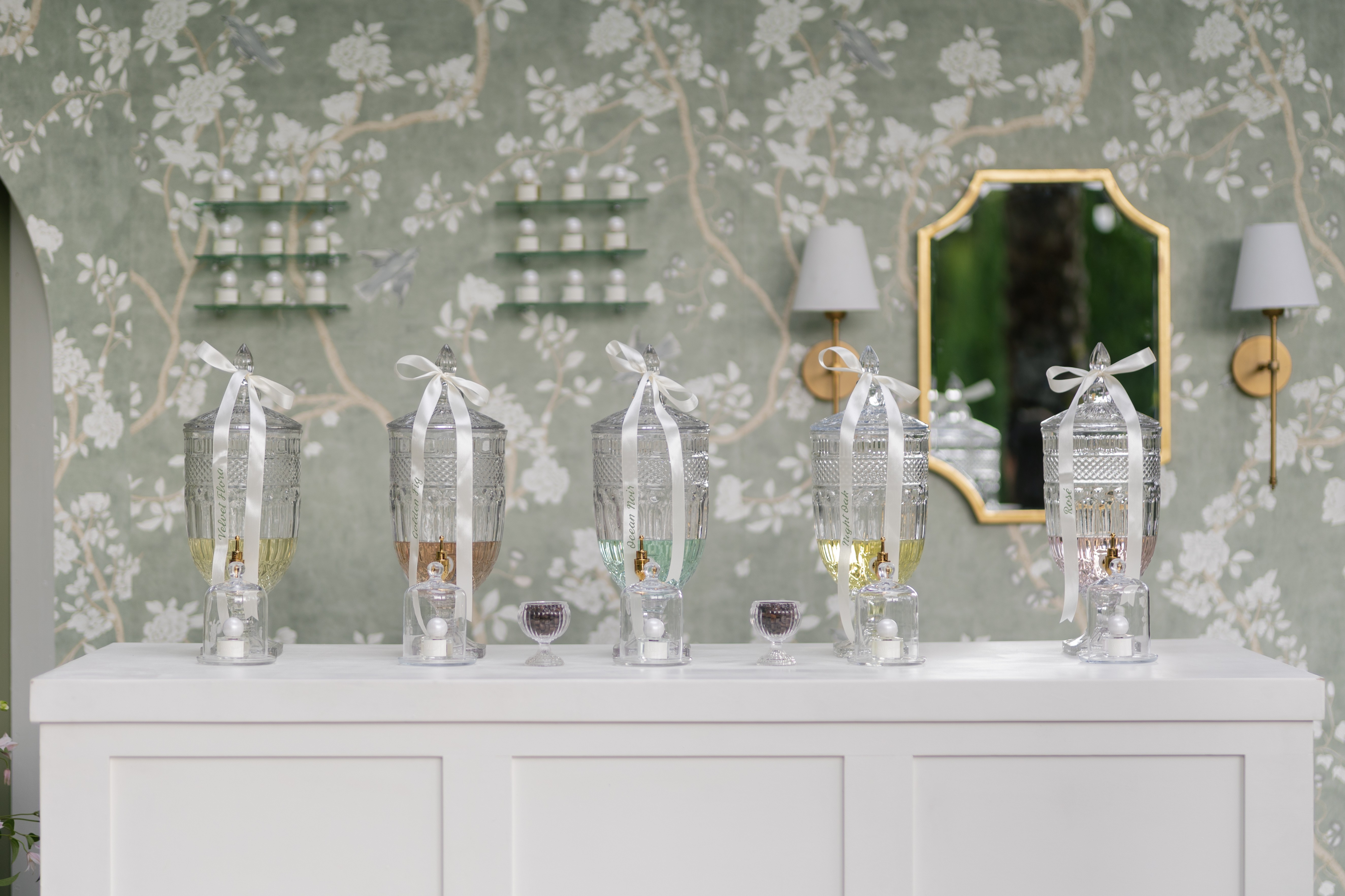 Decorative wall with floral pattern, white sideboard with six water bottles, mirror, and wall-mounted lamps.