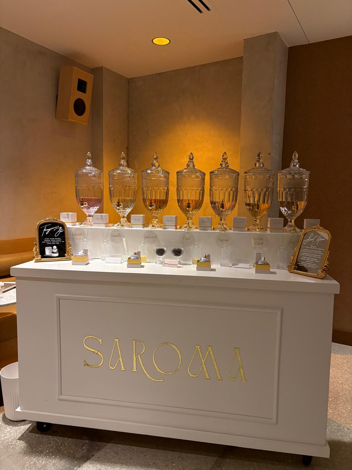 Display of seven large glass jars with lids on a white counter, with a sign reading 'SAROMA' in front.