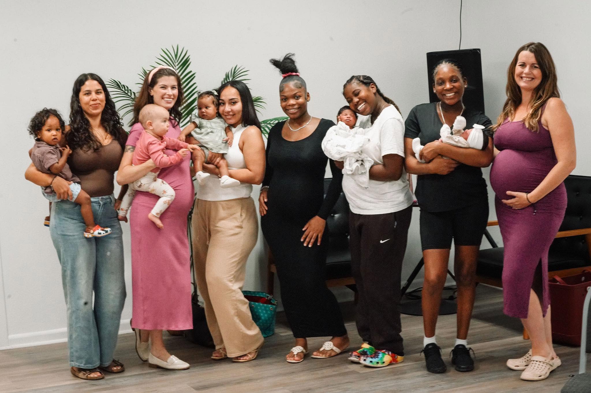 Group of women and children standing indoors, some holding babies, smiling, with plants and a blackboard in background.