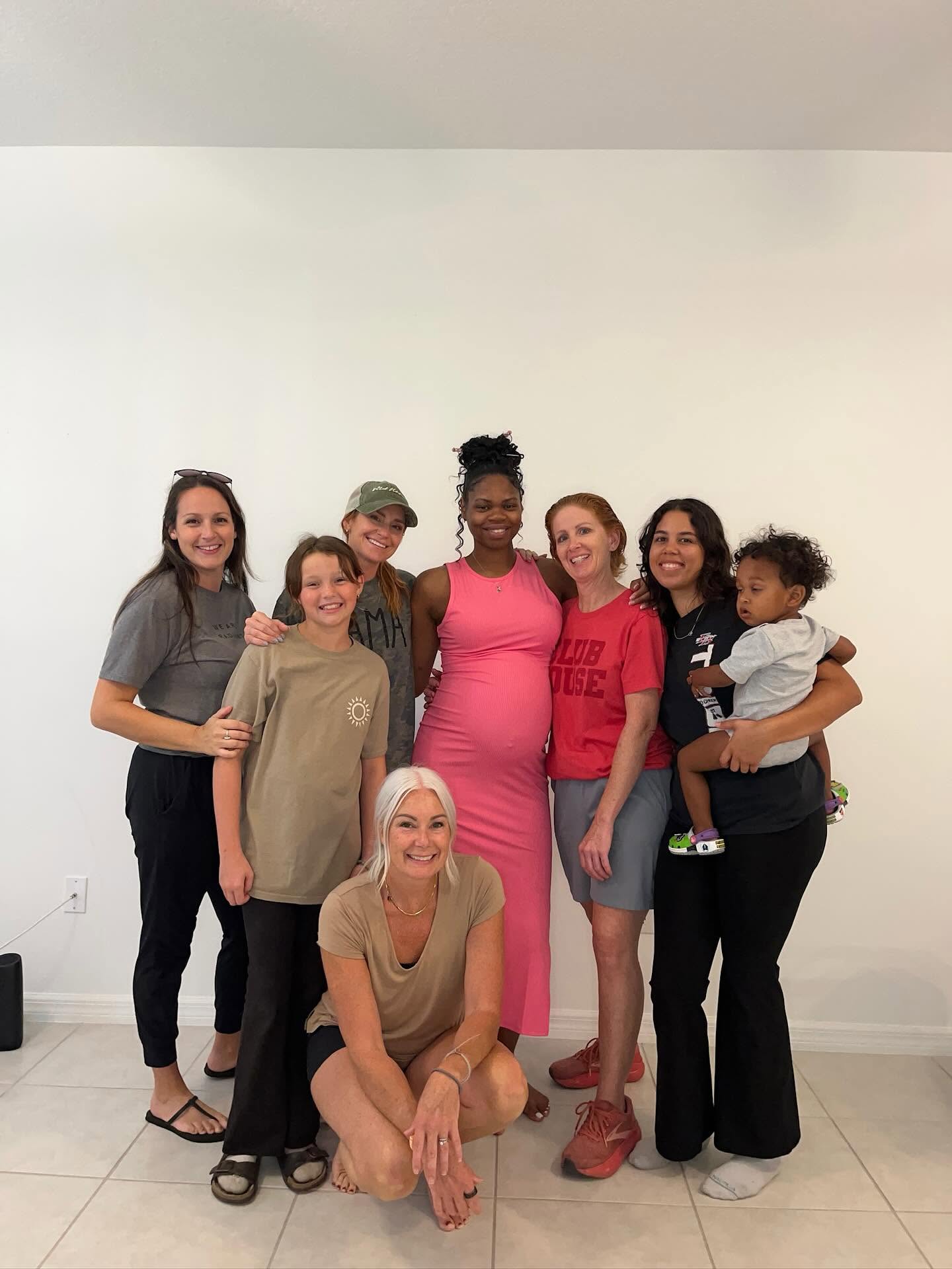 Seven women and a child standing together against a plain white wall, smiling, with one woman sitting on the floor.