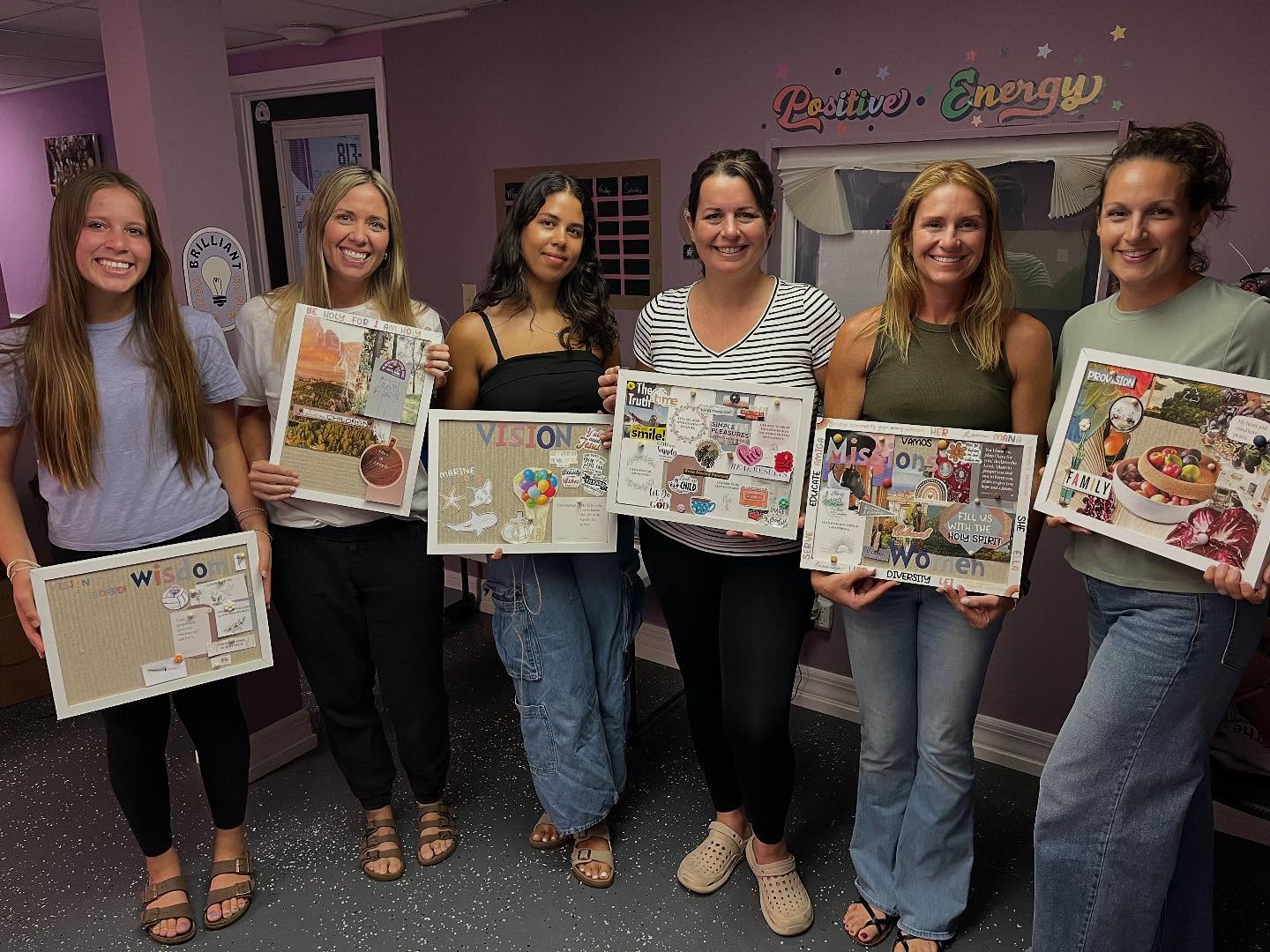 Six women standing indoors, holding colorful boxes and posters, smiling for the photo, with a purple wall and colorful sign in the background.