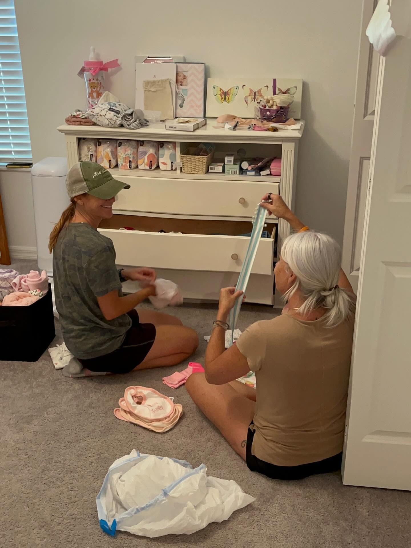 Two people sitting on the floor organizing items in a room, with a white dresser and various objects on top.