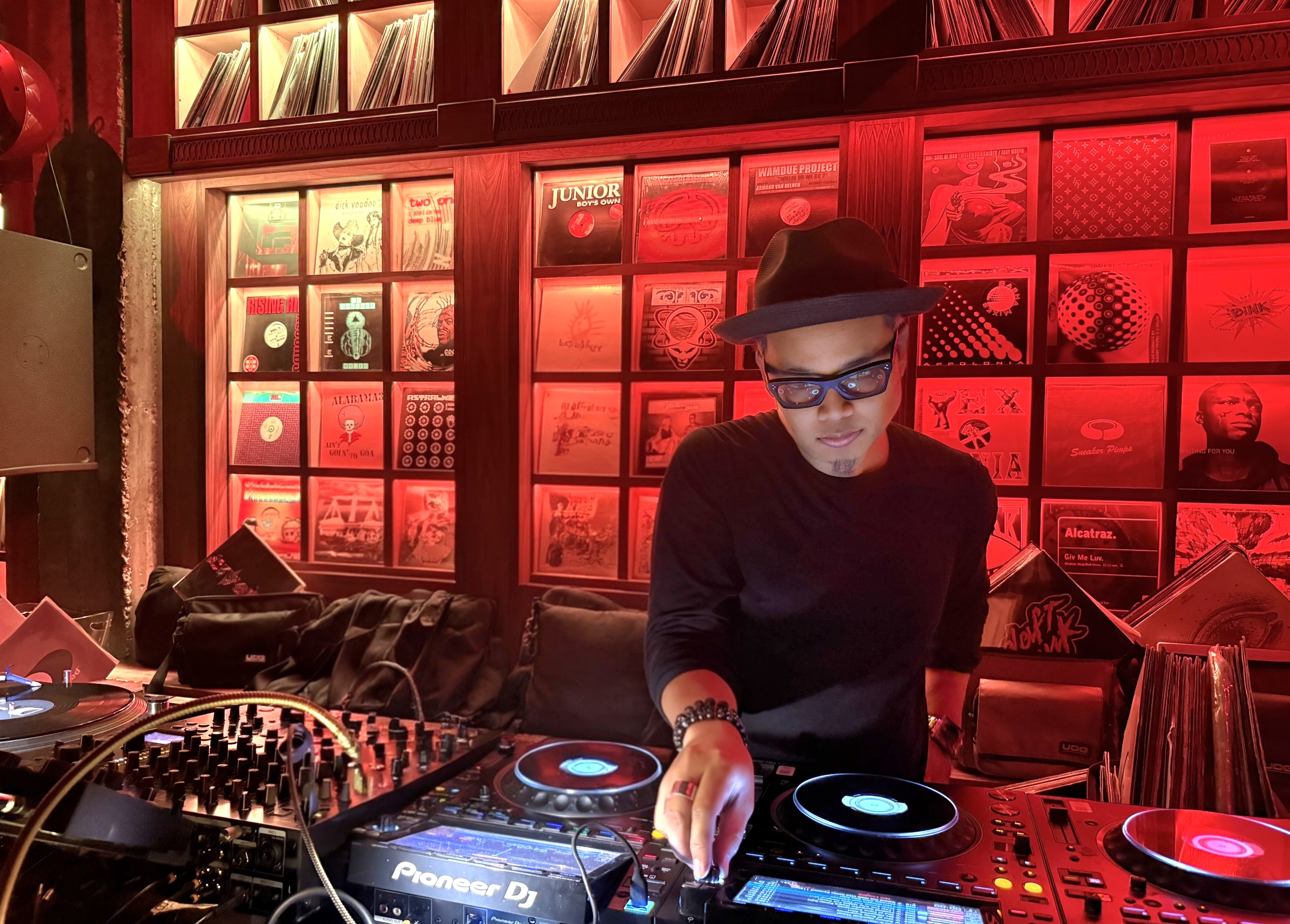 DJ wearing glasses and a hat operates turntables in a red-lit room with wall art, equipment, and a sofa.