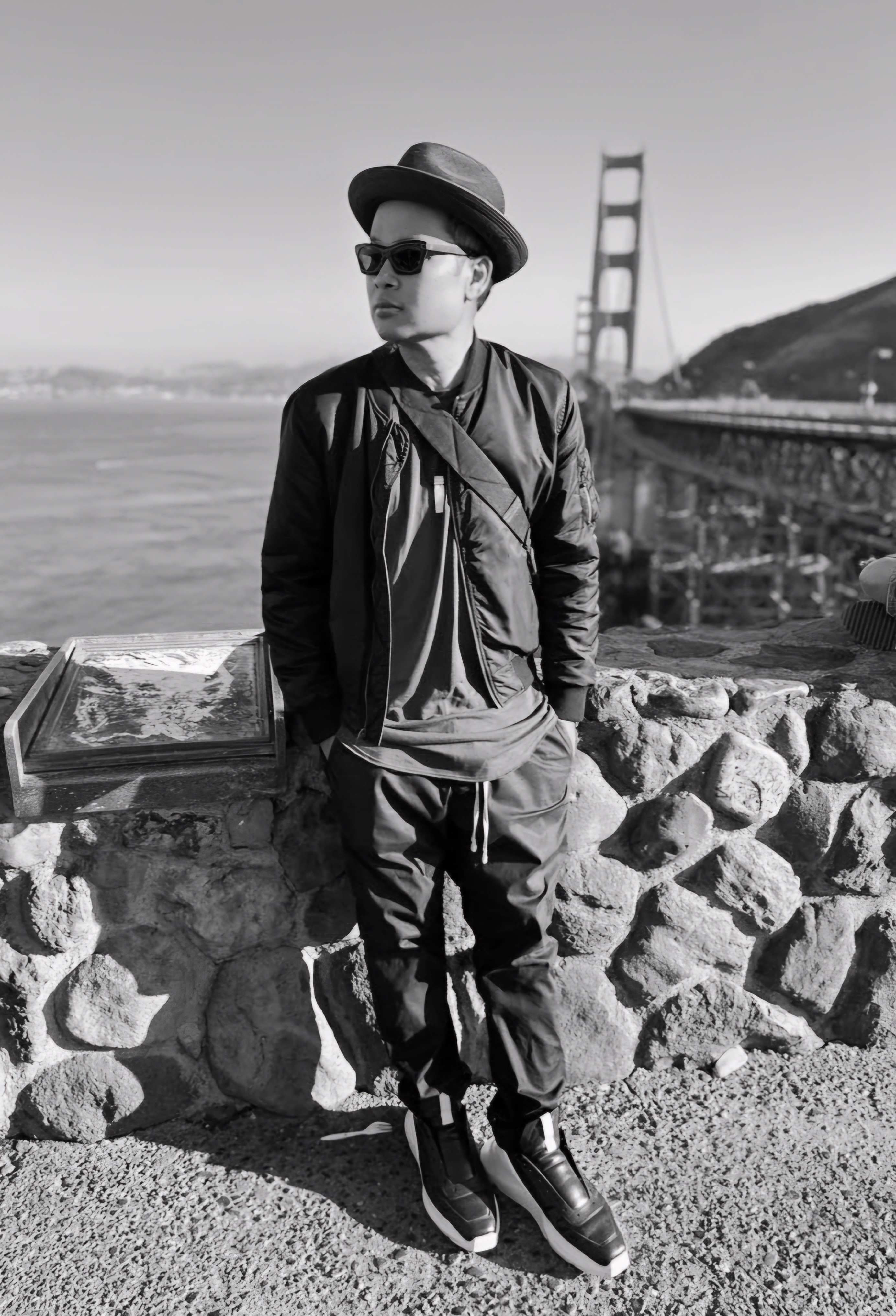 Person wearing sunglasses, hat, and jacket leaning against a stone wall near a bridge, with water and hills in background.
