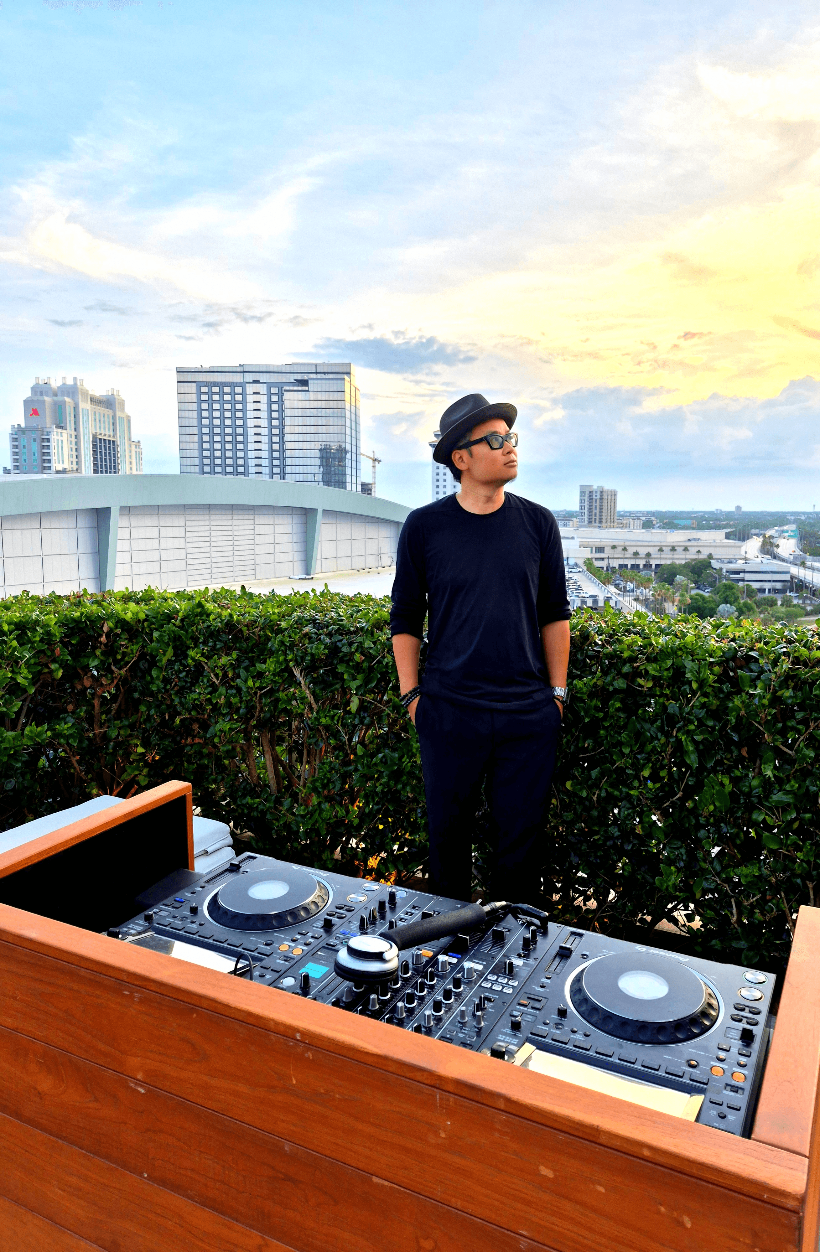 Person wearing a black hat and shirt stands outdoors with city buildings and sky in background.