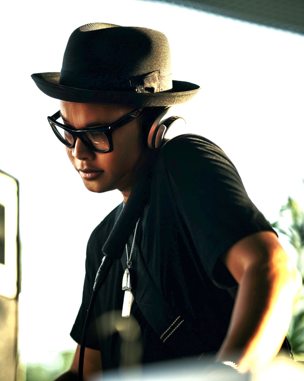 Person wearing a hat, glasses, and headphones, looking down, with a bright background and some greenery.