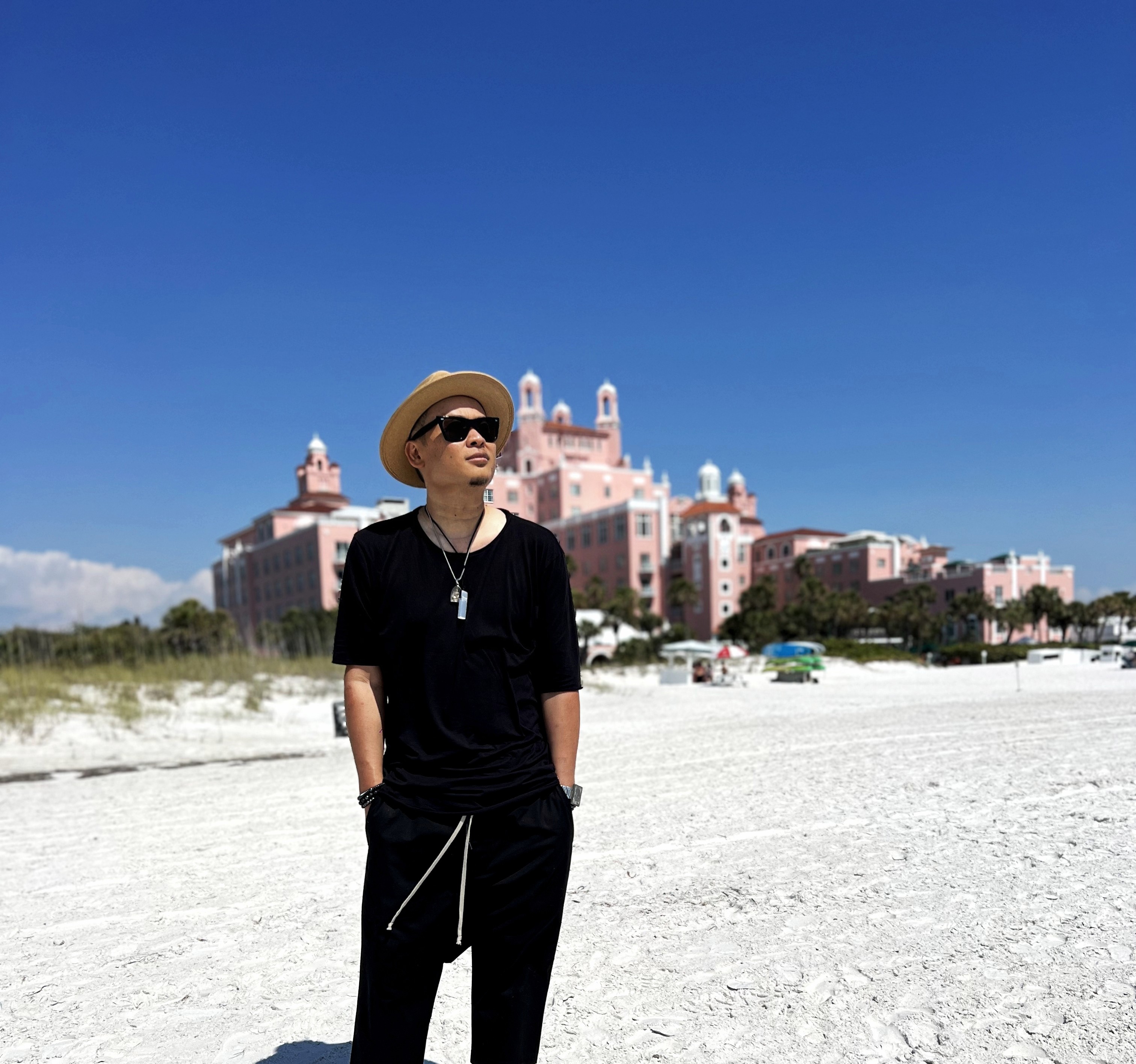 Person wearing sunglasses, hat, and black outfit standing on sandy beach with pink castle-like building in background under blue sky.