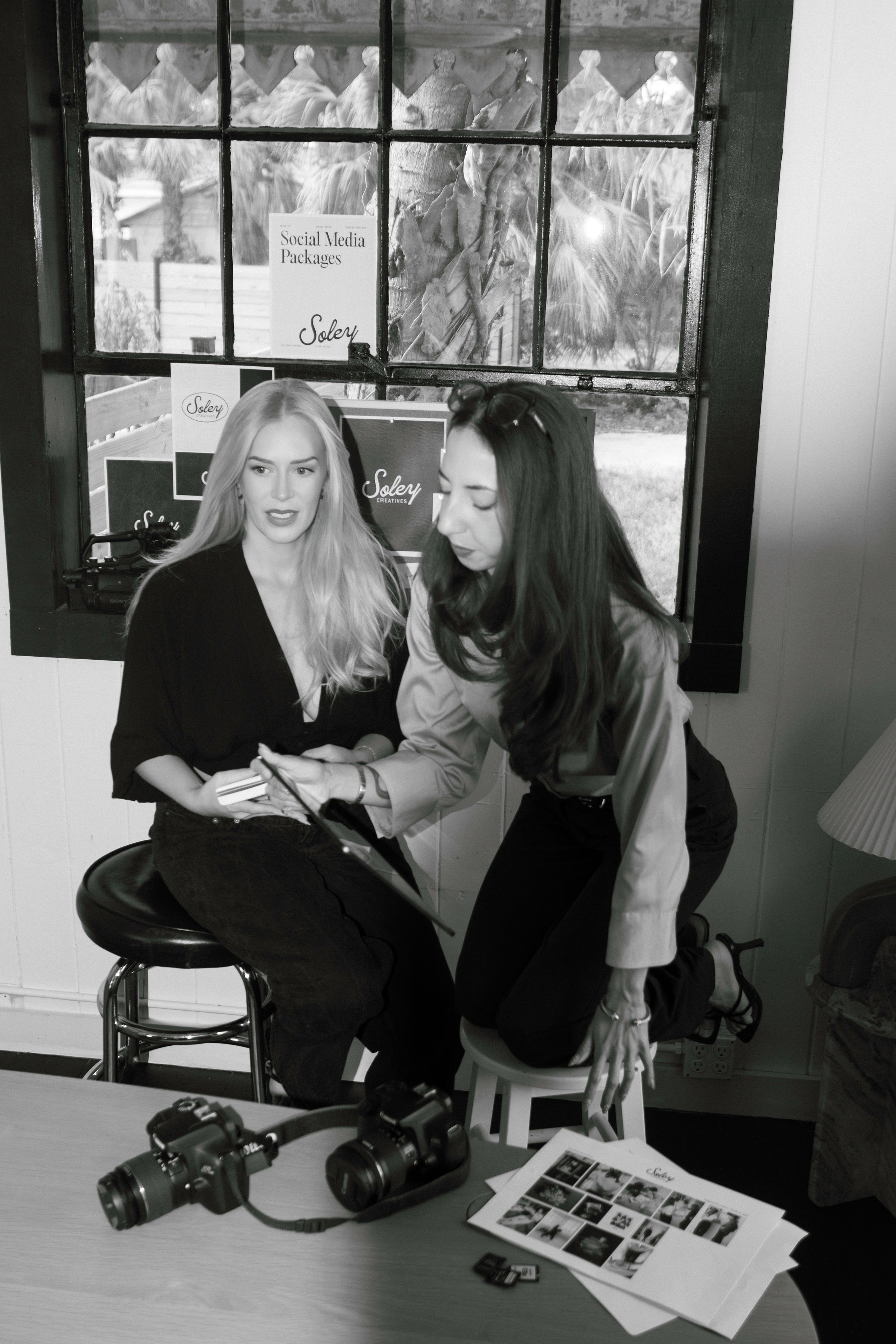 Two women sit at a table with cameras and printed photos, engaging in conversation indoors near a window.