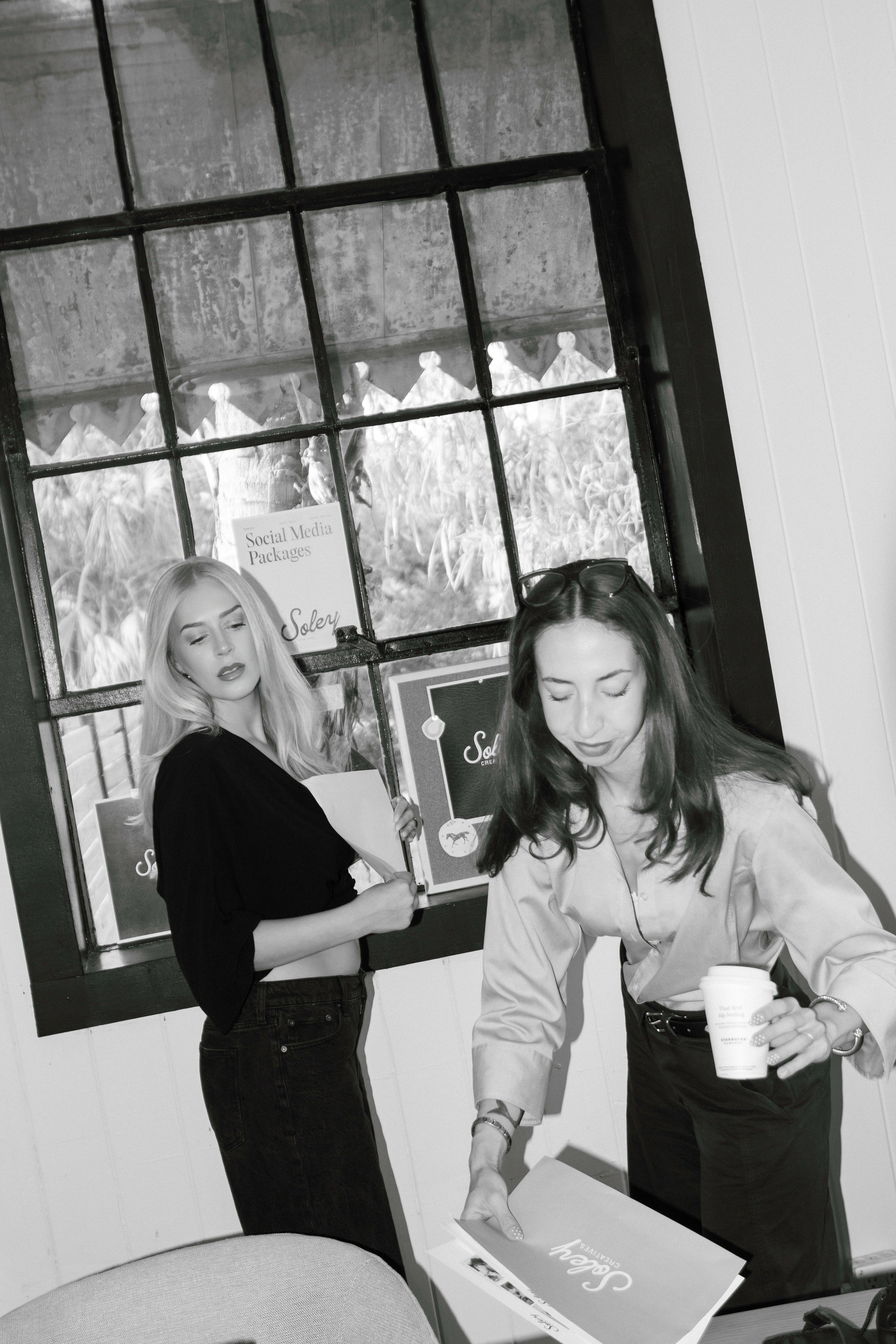 Two women near a large window, one holding a cup and a box, the other looking at her, in black and white.