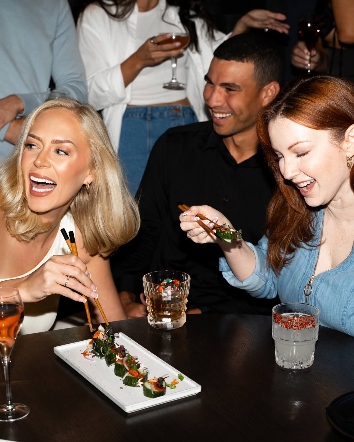 Three people smiling and laughing at a party, with drinks and a plate of food on the table.