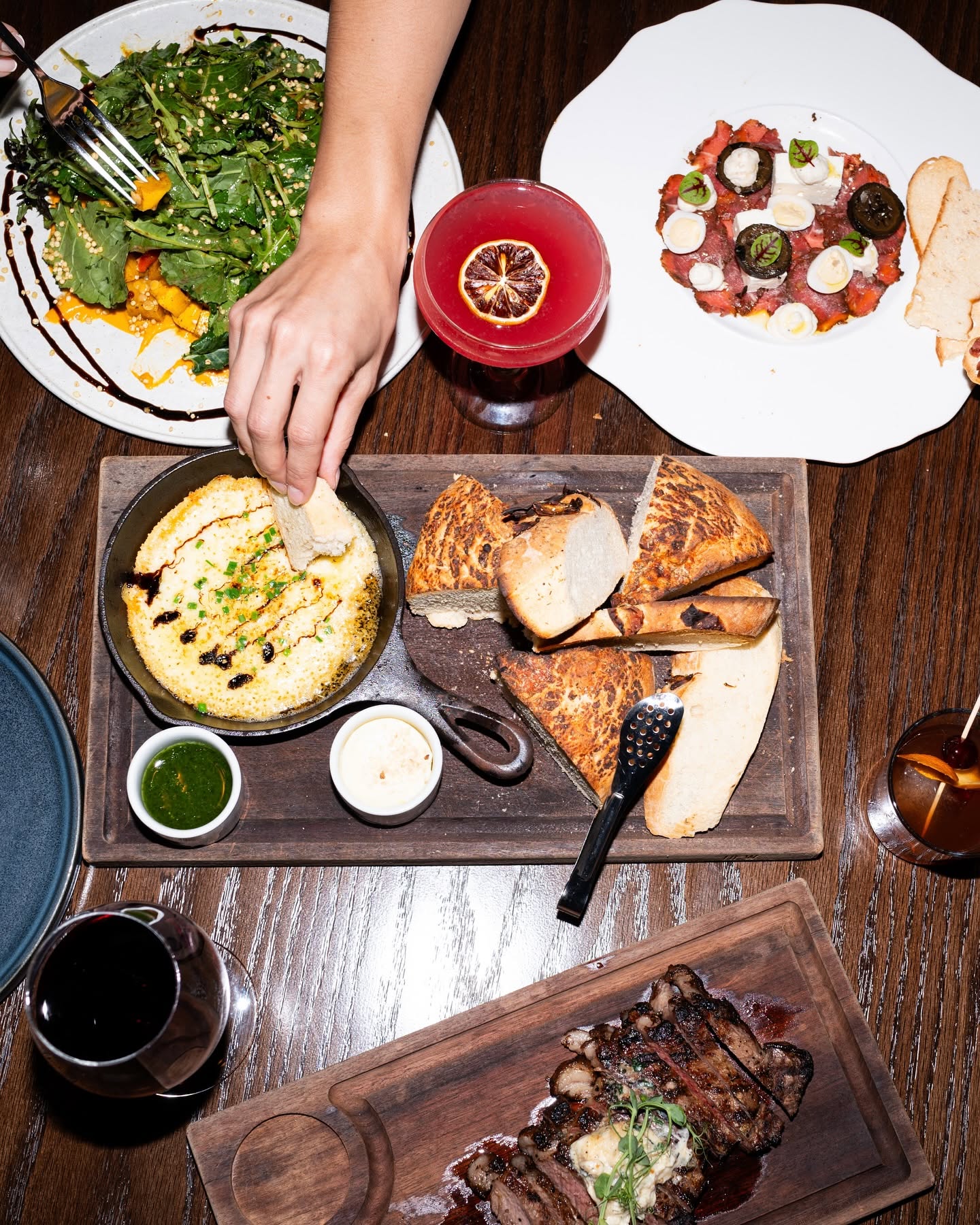 Person reaching for a dish with salad, two plates with appetizers, bread, and drinks on a wooden table.