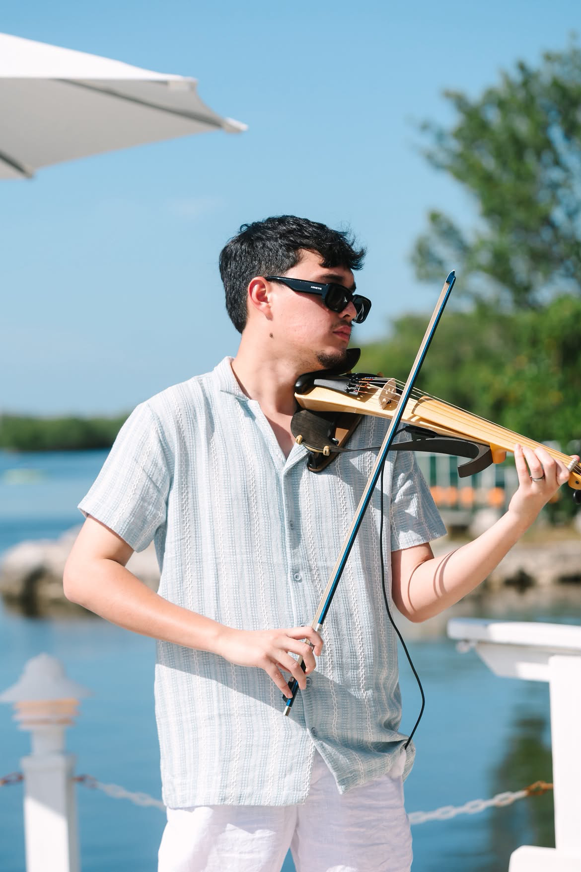 Young man playing violin outdoors near water, wearing sunglasses and a striped shirt, with trees and sky in background.