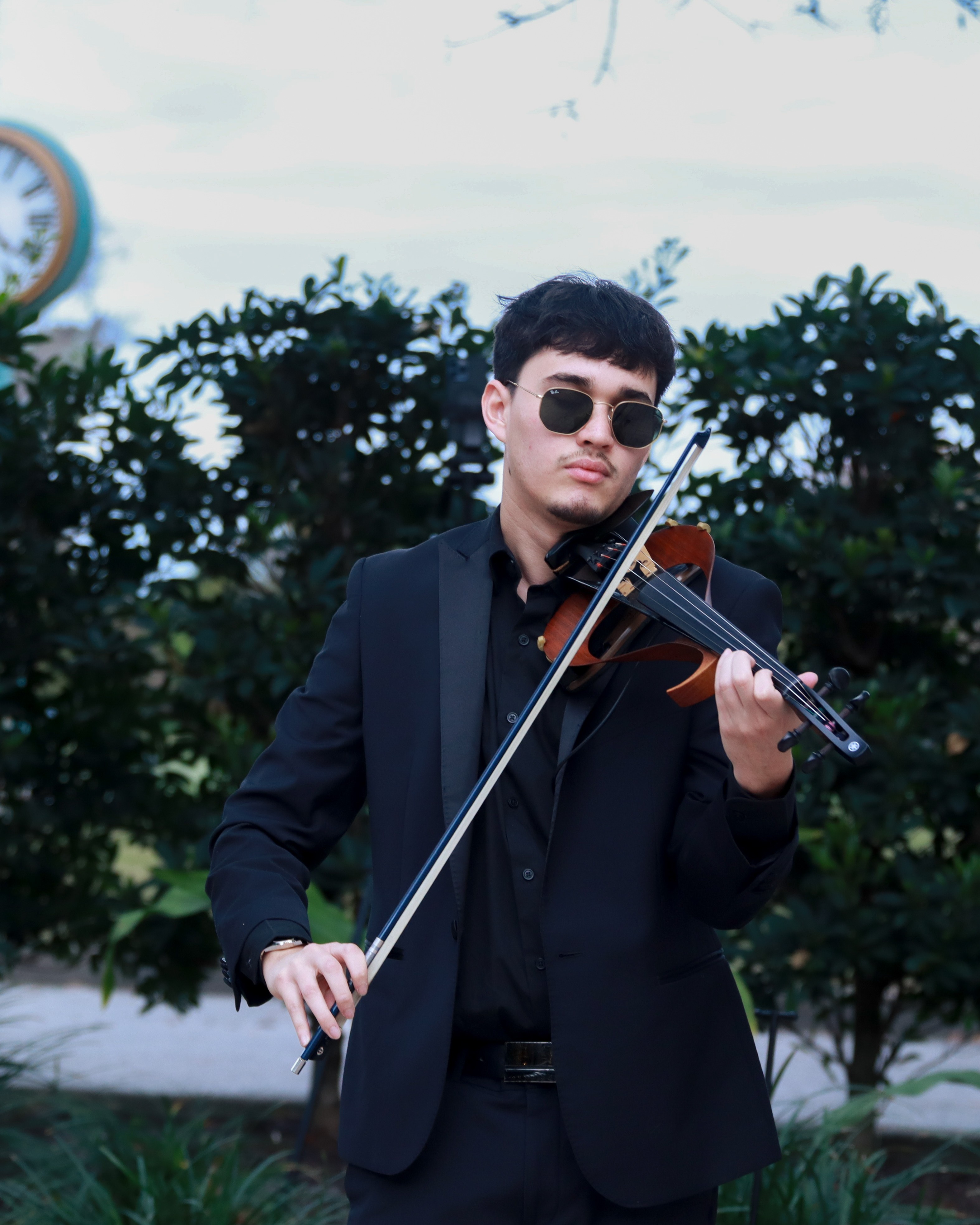 Young man in sunglasses playing violin outdoors with trees and sky in background.