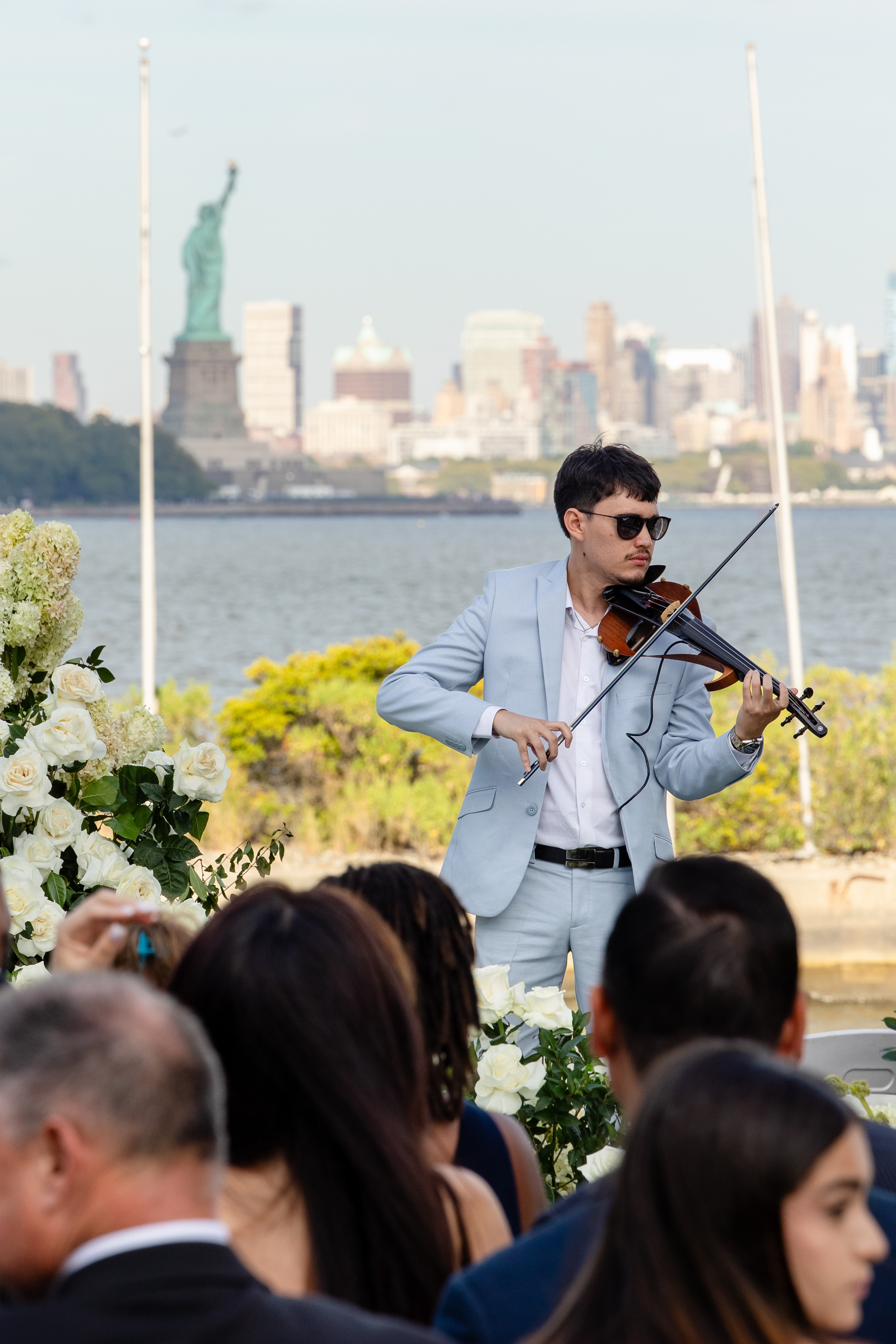 Man playing violin outdoors with city skyline and Statue of Liberty in background, audience in foreground.