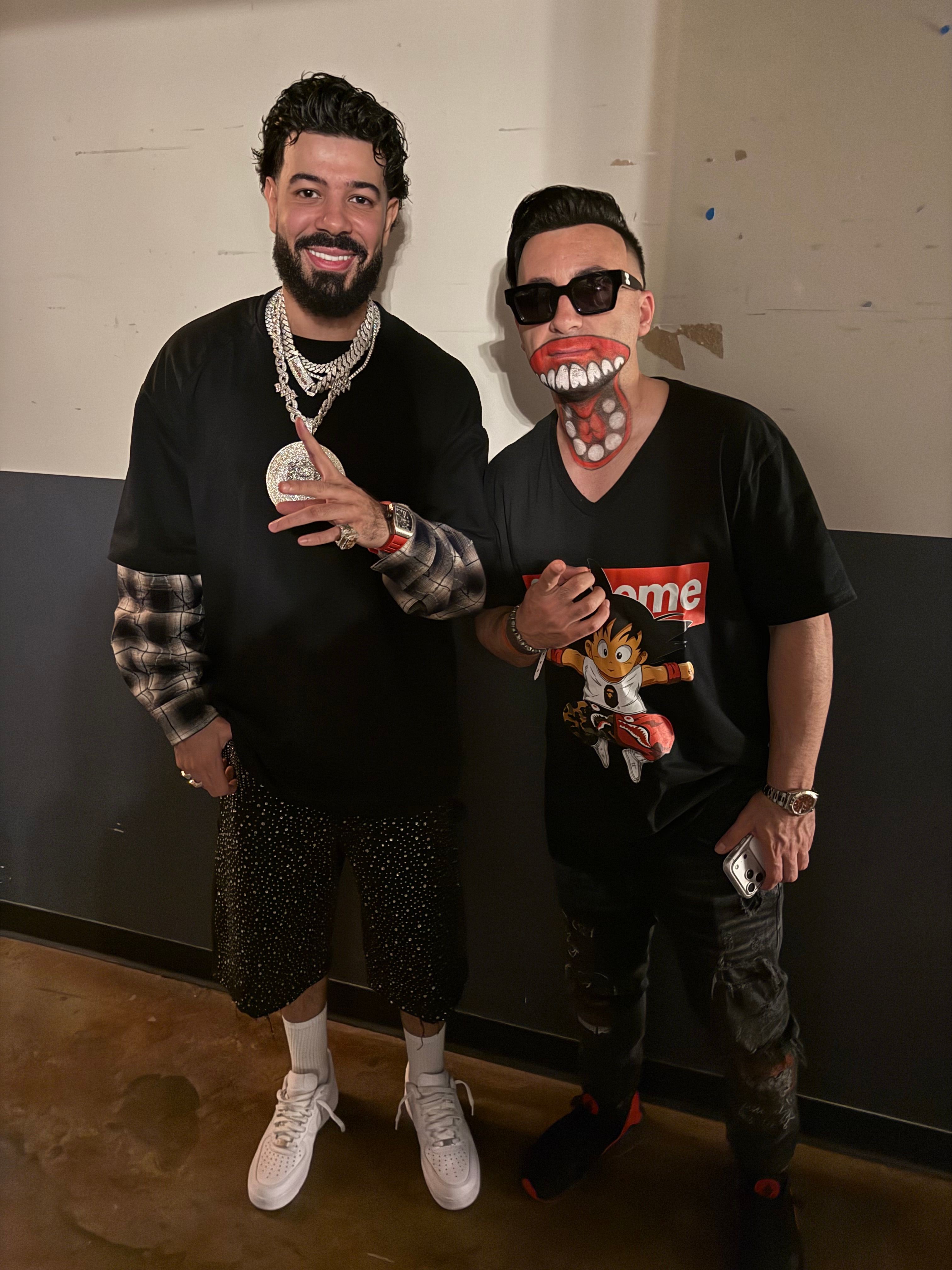 Two men dressed in black with accessories, one holding a small plush toy, standing indoors against a plain wall.