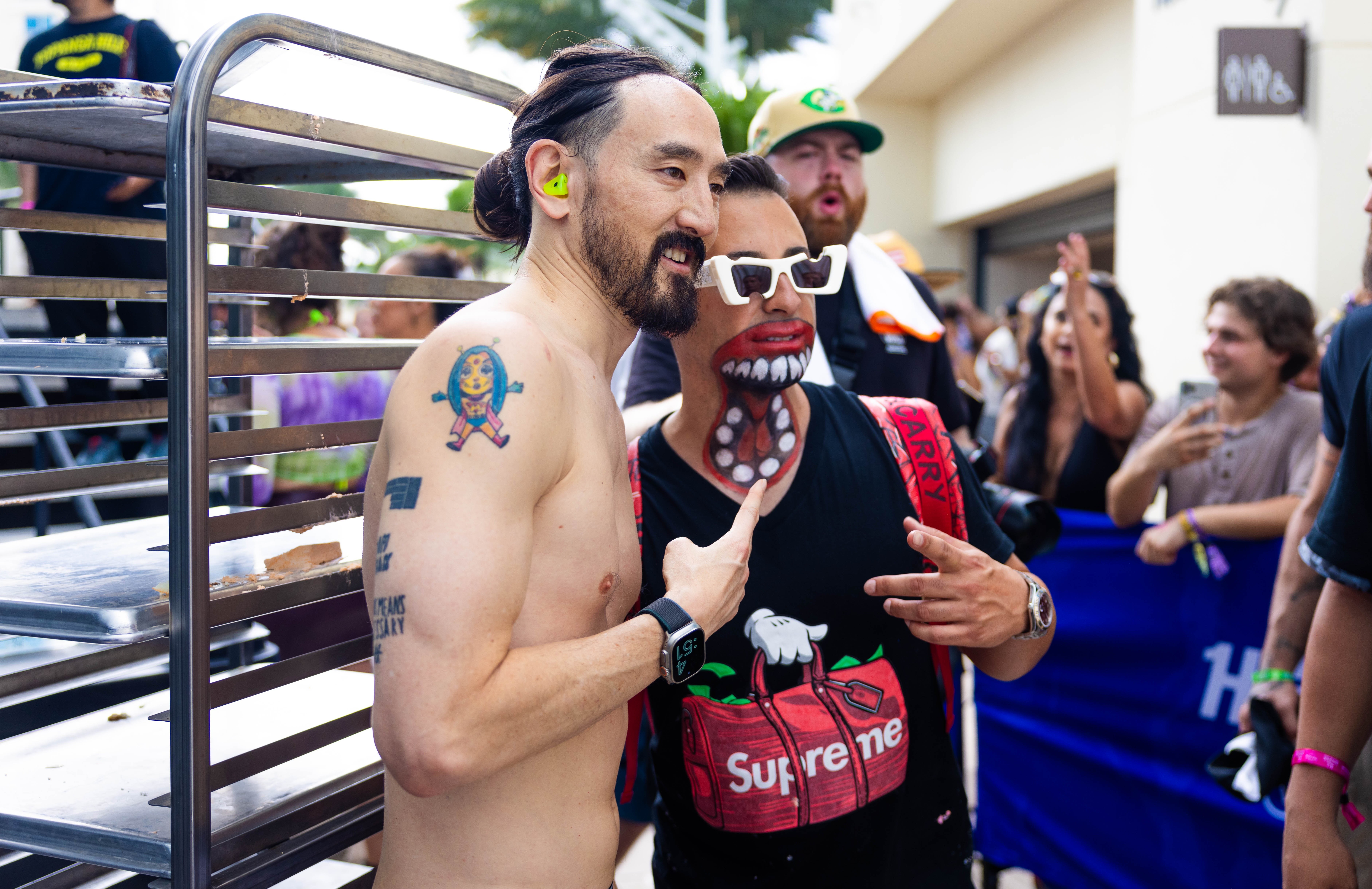 Two people at an outdoor event, one with tattoos and the other wearing a mask with a large mouth, surrounded by others.